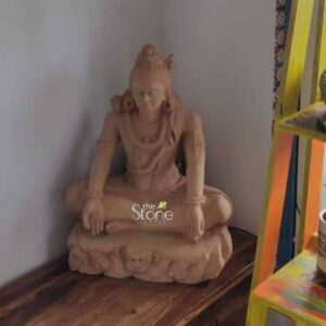 A serene 2-foot sandstone Shiva statue is elegantly displayed on a polished wooden surface in an indoor setting likely a home or meditation space in Bangalore. The statue depicts Lord Shiva in a meditative posture seated in Padmasana exuding an aura of profound tranquility and divine wisdom. The natural light brown hue of the sandstone allows the intricate carvings to be subtly highlighted contributing to a calm and spiritual ambiance. Lord Shiva's features are sculpted with a gentle and contemplative expression his eyes likely downcast in meditation though not clearly visible from this angle. His long flowing matted hair is piled high adorned with traditional elements. He wears simple necklaces and armlets carved with understated elegance. His hands rest gently in his lap in a meditative mudra. The base of the statue is ornately carved with stylized figures possibly skulls or mythical creatures adding a traditional and symbolic foundation to the divine figure. In the background a light-colored wall and a colorful multi-tiered shelf with decorative items suggest a thoughtfully curated interior. This 2-foot sandstone Shiva statue is an exceptional piece that brings an immediate sense of calm and spiritual harmony to any environment. Its natural sandstone material and meditative depiction make it a versatile addition to various decor styles particularly suited for a home in Bangalore. As a symbol of peace destruction of ignorance and cosmic creation this Shiva statue is not only an artistic masterpiece but also a significant spiritual icon inviting contemplation and fostering a tranquil atmosphere for reflection and peace.
