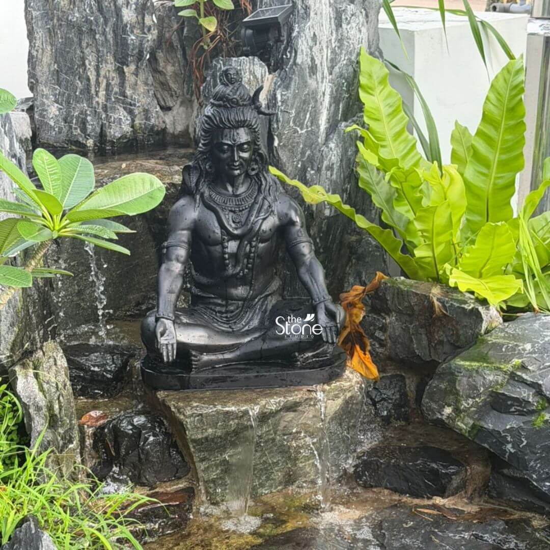 A majestic 2-foot black granite Lord Shiva statue is perfectly integrated into a naturalistic rock waterfall feature in an outdoor garden creating a serene and spiritual focal point. The statue depicts Lord Shiva in a traditional meditative posture seated in Padmasana, exuding an aura of profound peace and power. The dark polished surface of the black granite contrasts beautifully with the verdant green foliage and the dark textured rocks making it a prominent and calming presence in this Iskandar Puteri Malaysia garden setting. Lord Shiva is adorned with his characteristic iconography matted hair piled high on his head a crescent moon and possibly the third eye subtly indicated on his forehead. He wears necklaces and armlets all carved with intricate detail into the hard granite. Water gently flows over the rocks beneath and around the statue creating a soothing natural soundscape and visually connecting Shiva to the element of water often associated with his sacred form. Lush tropical plants including broad-leafed ferns and smaller shrub frame the statue enhancing the natural and sacred ambiance. This 2-foot Black Granite Shiva Statue is an exceptional piece that transforms the garden into a sanctuary for meditation and spiritual reflection. Its durable granite construction ensures longevity in outdoor conditions while its timeless design makes it a versatile element in various garden styles. Beyond its aesthetic appeal this Shiva statue serves as a powerful symbol of tranquility destruction of ignorance and cosmic creation making it a deeply meaningful and revered addition to any outdoor space particularly in a tropical setting like Malaysia.