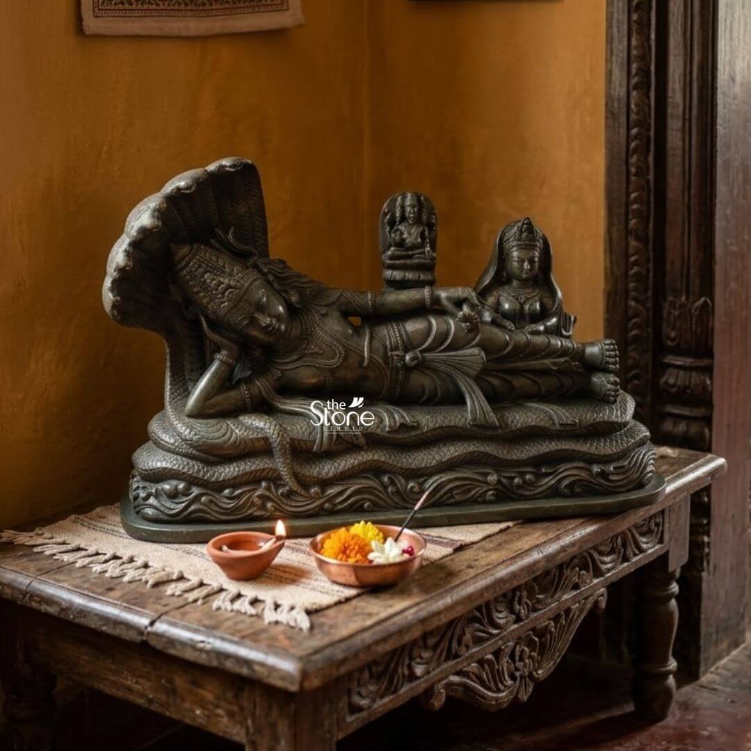 Ananta Shayana Vishnu Statue 20" 3 A profound green stone statue of Lord Vishnu in his iconic sleeping posture is prominently displayed on a richly carved wooden table set against an earthy-toned wall. The statue captures Vishnu reclining on the cosmic serpent Sheshnag symbolizing the universe's cyclical nature of dissolution and creation. The deep verdant green of the stone possibly a dark serpentine or steatite provides a majestic and ancient aestheti highlighting the intricate details of the figures and the serene spiritual narrative making it an ideal idol for a temple setting. Lord Vishnu is depicted in a state of serene cosmic slumber his head gently supported by his right arm with an expression of ultimate tranquility. He is lavishly adorned with traditional Hindu ornaments including an elaborate crown multiple necklaces, intricate armlets, and a gracefully draped dhoti all carved with meticulous precision. The multi-headed Sheshnag coils beneath him its numerous hoods fanning out to form a grand protective canopy over Vishnu's head. From Vishnu's navel a delicate lotus emerges bearing a miniature figure of Brahma the creator god. To Vishnu's right seated attentively is Goddess Lakshmi his divine consort her form equally adorned and sculpted with reverence attending to his feet. In the foreground on the table a small lit oil lamp and a copper bowl with flowers serve as traditional offerings. This green stone Sleeping Vishnu idol is a powerful and sacred representation of preservation cosmic order and the continuous cycle of time perfectly suited for a temple. The exceptional detail and the choice of green stone often associated with spiritual balance and renewal significantly enhance its profound symbolism. This exquisitely crafted statue surrounded by traditional offerings creates a deeply spiritual ambiance inviting contemplation and reverence for the divine cosmic dance and enriching any temple or sacred space with its deep cultural and religious resonance.