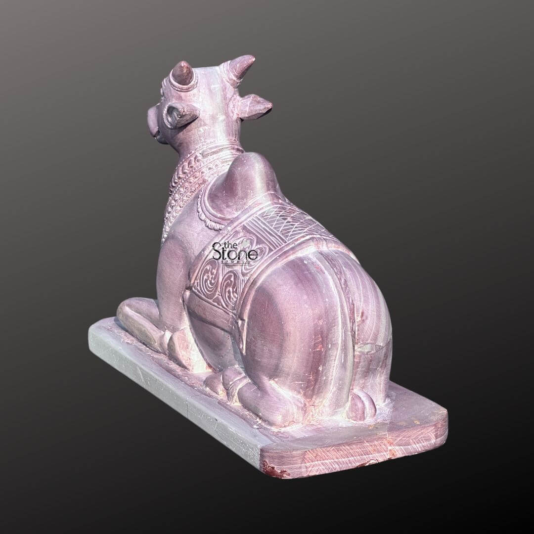 Stone Nandi Statue for Home Temple 8" 6 A beautifully crafted 8-inch Nandi Murti sculpted from natural pink stone is presented in a slightly elevated rear-left view against a muted background. The statue captures the revered bull Nandi in a traditional seated pose embodying devotion and strength. The subtle natural pink variations in the stone add warmth and character to the piece highlighting the smooth polished surfaces and the artisan's attention to detail making it an ideal spiritual accent for any sacred space. The Nandi's back is adorned with intricate carvings resembling a decorative blanket featuring symmetrical patterns and flowing designs that enhance the statue's visual appeal and cultural significance. The distinct hump of the sacred bull is prominently depicted and the contours of its powerful hindquarters are rendered with a gentle realism. The statue rests firmly on a minimalist rectangular base providing a stable foundation and emphasizing its role as a devotional object. This exquisite small Nandi Murti carved from natural pink stone serves as a powerful symbol of loyalty and purity perfectly aligning with its use in spiritual practices and home altars. Its compact 8-inch size makes it versatile for various settings while the natural pink stone imbues it with a gentle serene energy. The detailed craftsmanship and the inherent beauty of the stone combine to create a deeply meaningful and aesthetically pleasing representation of Lord Shiva's divine vehicle fostering an atmosphere of peace and reverence.