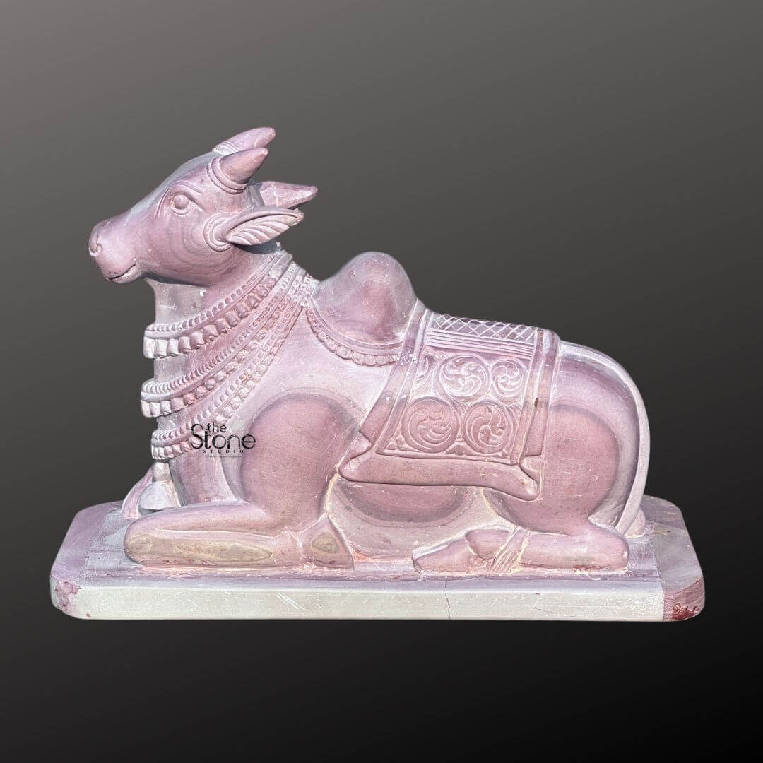 Stone Nandi Statue for Home Temple 8" 7 A beautifully carved 8-inch Nandi statue crafted from pink stone is shown in a profile view against a plain dark background. The statue depicts Nandi the sacred bull in a serene reclining position with its head held high and turned slightly towards the left. The natural pink coloration of the stone is evident showcasing varying shades and textures that highlight the contours of the sculpture and the meticulous details of its design making it a compelling piece for spiritual or decorative purposes. The Nandi is adorned with intricately carved decorative elements including a multi-layered necklace around its neck and an ornate saddle or blanket draped over its back. These embellishments feature delicate patterns swirled motifs and geometric designs adding a rich cultural and artistic dimension to the statue. The bull's characteristic hump is prominently sculpted and its powerful yet calm physique is rendered with flowing lines that convey both strength and tranquility. This small-sized 8-inch Nandi statue in pink stone is a striking representation of devotion and purity perfectly embodying the essence of its role in Hindu mythology. Its compact dimensions make it suitable for a variety of settings such as a home altar meditation space or as an elegant piece of decor. The thoughtful craftsmanship and the gentle aesthetic of the pink stone make this Nandi statue a meaningful and visually appealing artifact that brings a touch of sacred beauty to any environment.