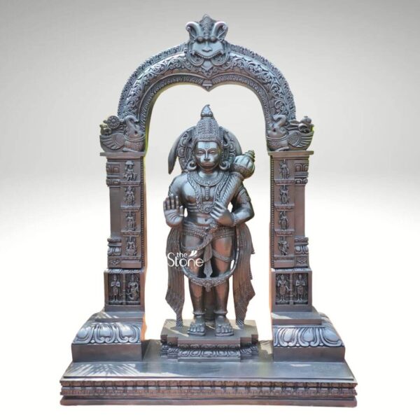 An imposing black stone Hanuman statue depicted as standing Bajrangbali is framed by an intricately carved temple archway presented against a plain light background. This stunning black stone statue captures Lord Hanuman in a powerful and dynamic standing posture exuding an aura of immense strength devotion and courage. The dark polished surface of the stone provides a dramatic contrast enhancing the intricate details of the sculpture and its elaborate architectural frame making it a powerful focal point for worship and admiration. Lord Hanuman is depicted with his characteristic features including a majestic crown prominent ear ornaments and a strong muscular physique. His right hand is raised in the Abhaya Mudra offering blessings and protection while his left hand holds his Gada symbolizing his immense power and readiness to vanquish evil. A garland of beads adorns his neck and a decorative sash drapes across his body. He stands on a lotus pedestal which is integrated into a multi-tiered base adorned with smaller figures and decorative carvings all contributing to the grandeur of the piece. The intricate temple archway above him is equally detailed featuring divine figures floral motifs and a Kirtimukha at the apex flanked by mythical creatures. This black stone Standing Bajrangbali statue is a profound representation of Lord Hanuman the epitome of strength devotion and selfless service. The exceptional craftsmanship in black stone not only makes it a visually striking piece but also embodies deep cultural and religious significance offering a powerful focal point for devotion and prayer. The rich detailing of the deity and his surrounding archway reflects centuries of tradition making it a truly remarkable and revered artifact that brings blessings protection and a spiritual presence into any sacred space.