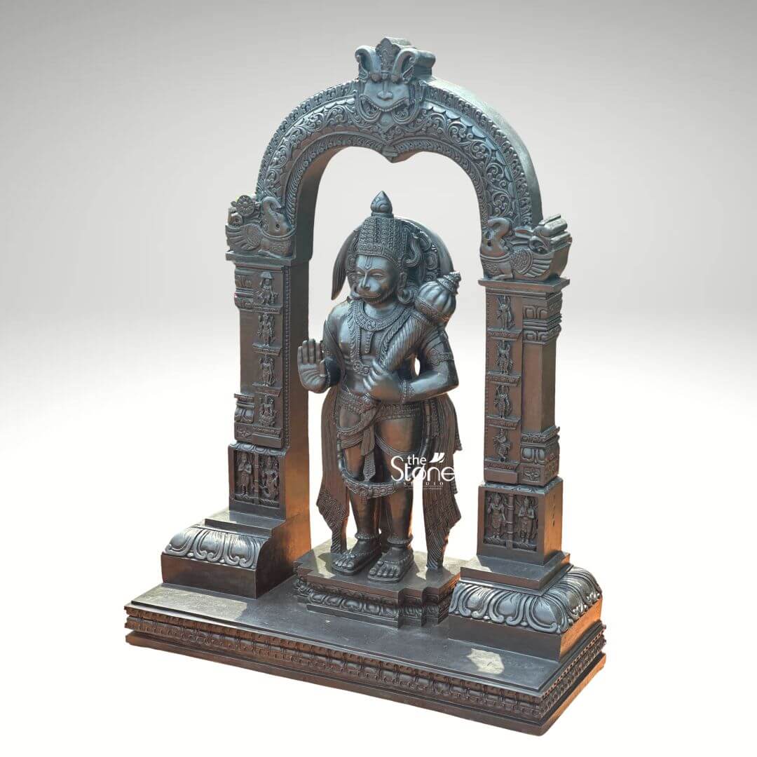 Standing Bajrangbali Statue 4ft 2 An imposing black stone Hanuman statue depicted as standing Bajrangbali within an ornate carved arch is presented against a plain light background. This stunning black stone statue captures Lord Hanuman in a powerful and dynamic standing posture exuding an aura of immense strength devotion and courage. The dark polished surface of the stone provides a dramatic contrast enhancing the intricate details of the sculpture and its elaborate architectural frame making it a powerful focal point for worship and admiration. Lord Hanuman is depicted with his characteristic features including a majestic crown prominent ear ornaments and a strong muscular physique that symbolizes his extraordinary power. His right hand is raised in the Abhaya Mudra offering blessings and protection to devotees while his left hand holds his Gada resting on his shoulder symbolizing his immense power and readiness to vanquish evil. A garland of beads adorns his neck and a decorative sash drapes across his body all carved with meticulous precision. He stands on a lotus pedestal which is integrated into a multi-tiered base adorned with smaller figures and decorative carvings all contributing to the grandeur of the piece. The intricately carved archway above him is equally detailed featuring divine figures floral motifs and a Kirtimukha at the apex flanked by mythical creatures. This black stone Standing Bajrangbali statue with its ornate carved arch is a profound representation of Lord Hanuman the epitome of strength devotion and selfless service. The exceptional craftsmanship in black stone not only makes it a visually striking piece but also embodies deep cultural and religious significance offering a powerful focal point for devotion and prayer. The rich detailing of the deity and his surrounding archway reflects centuries of tradition making it a truly remarkable and revered artifact that brings blessings protection and a spiritual presence into any sacred space.