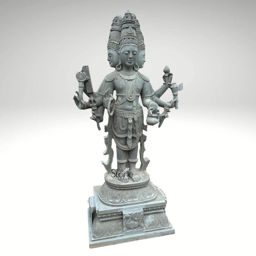 Brahma Stone Statue 7ft 10 This full-length seven-foot-tall standing statue of Lord Brahma carved from grey stone is presented against a plain white background allowing its intricate details to stand out. The deity is depicted with his characteristic four serene faces each adorned with a tall, ornate crown and looking in a different direction symbolizing his omnipresence. Brahma stands tall on a multi-tiered intricately carved pedestal showcasing his divine posture and the traditional iconography associated with the creator god. His numerous arms extend gracefully each hand holding a symbolic object. The craftsmanship of this grey stone Brahma statue is evident in the fine details etched into the stone. Brahma's hands hold essential items such as the kamandalu a mala a pustaka and a sruk all significant to his role as the creator and patron of knowledge. The sculptor has meticulously rendered the deity's jewelry including necklaces armbands and anklets, as well as the flowing drapes of his dhoti adding depth and realism to the stone figure. The grey stone material lends an air of timelessness and solemnity to the statue. As a central image for an article on Standing Brahma Stone Statues this seven-foot greystone rendition is a perfect example of traditional Hindu artistry. The statue's standing pose along with its detailed base emphasizes its stature and significance in a temple or devotional setting. The overall composition and the careful rendering of each feature make this a truly majestic representation of Lord Brahma embodying creation, wisdom, and the enduring power of grey stone sculpture.