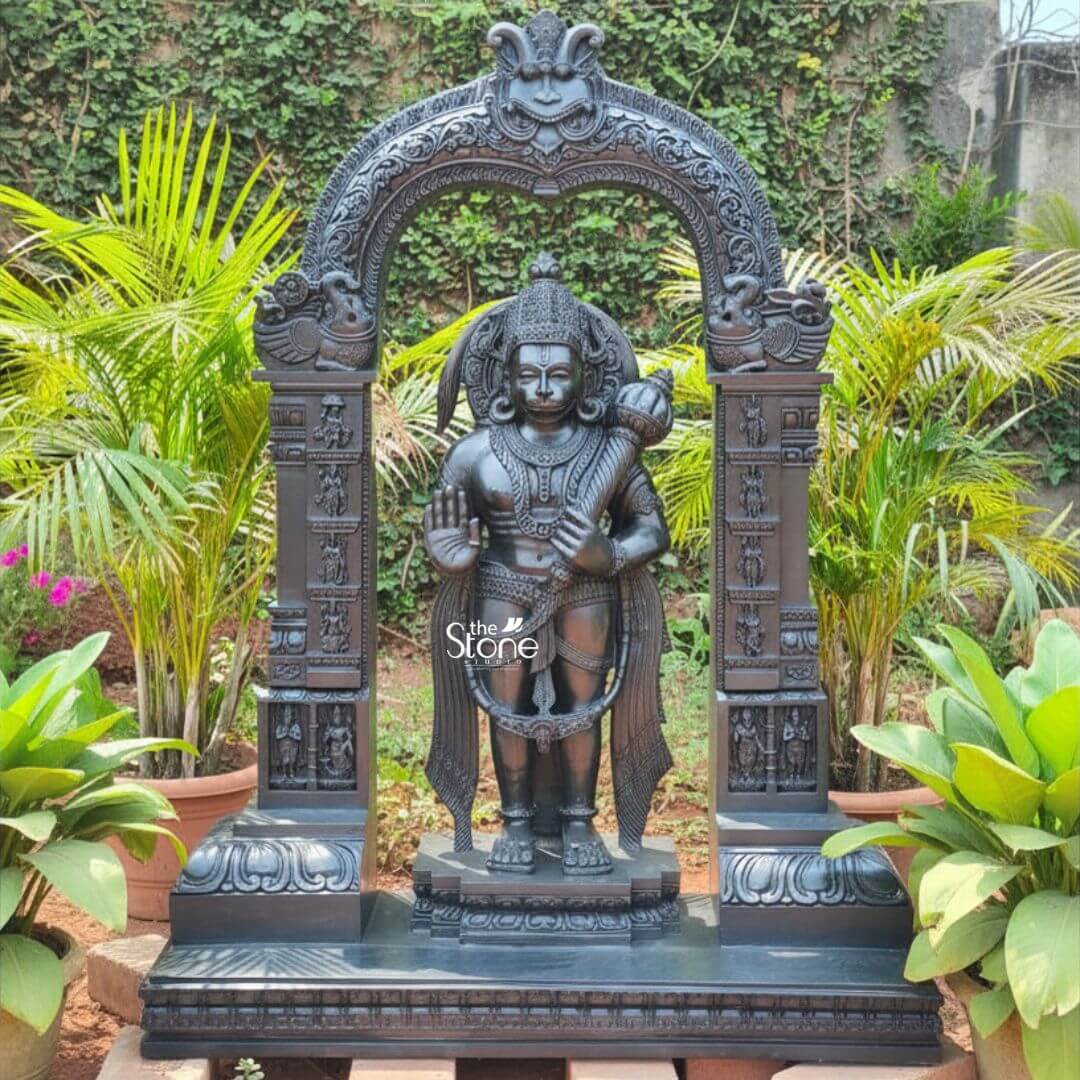 Standing Bajrangbali Statue 4ft 3 An impressive black stone Standing Hanuman Bhagwan Murti framed by an intricately carved temple arch is showcased outdoors against a lush green backdrop of potted plants and a creeper-covered wall. This stunning black stone statue captures Lord Hanuman in a powerful and dynamic standing posture exuding an aura of immense strength devotion and courage making it an ideal centerpiece for a mandir or a prominent garden display. The dark polished surface of the stone provides a dramatic contrast enhancing the intricate details of the sculpture and its elaborate architectural frame all within a vibrant natural setting. Lord Hanuman is depicted with his characteristic features including a majestic crown prominent ear ornaments and a strong muscular physique that symbolizes his extraordinary power. His right hand is raised in the Abhaya Mudra offering blessings and protection to devotees while his left hand holds his Gada resting on his shoulder symbolizing his immense power and readiness to vanquish evil. A garland of beads adorns his neck and a decorative sash drapes across his body all carved with meticulous precision. He stands on a lotus pedestal which is integrated into a multi-tiered base adorned with smaller figures and decorative carvings all contributing to the grandeur of the piece. The intricate temple archway above him is equally detailed featuring divine figures floral motifs and a Kirtimukha at the apex flanked by mythical creatures. This black stone Standing Hanuman Bhagwan Murti with its ornate carved arch is a profound representation of Lord Hanuman the epitome of strength devotion and selfless service. The exceptional craftsmanship in black stone not only makes it a visually striking piece but also embodies deep cultural and religious significance offering a powerful focal point for devotion and prayer in a mandir. The rich detailing of the deity and his surrounding archway reflects centuries of tradition making it a truly remarkable and revered artifact that brings blessings protection and a spiritual presence into any sacred space indoor or outdoor.