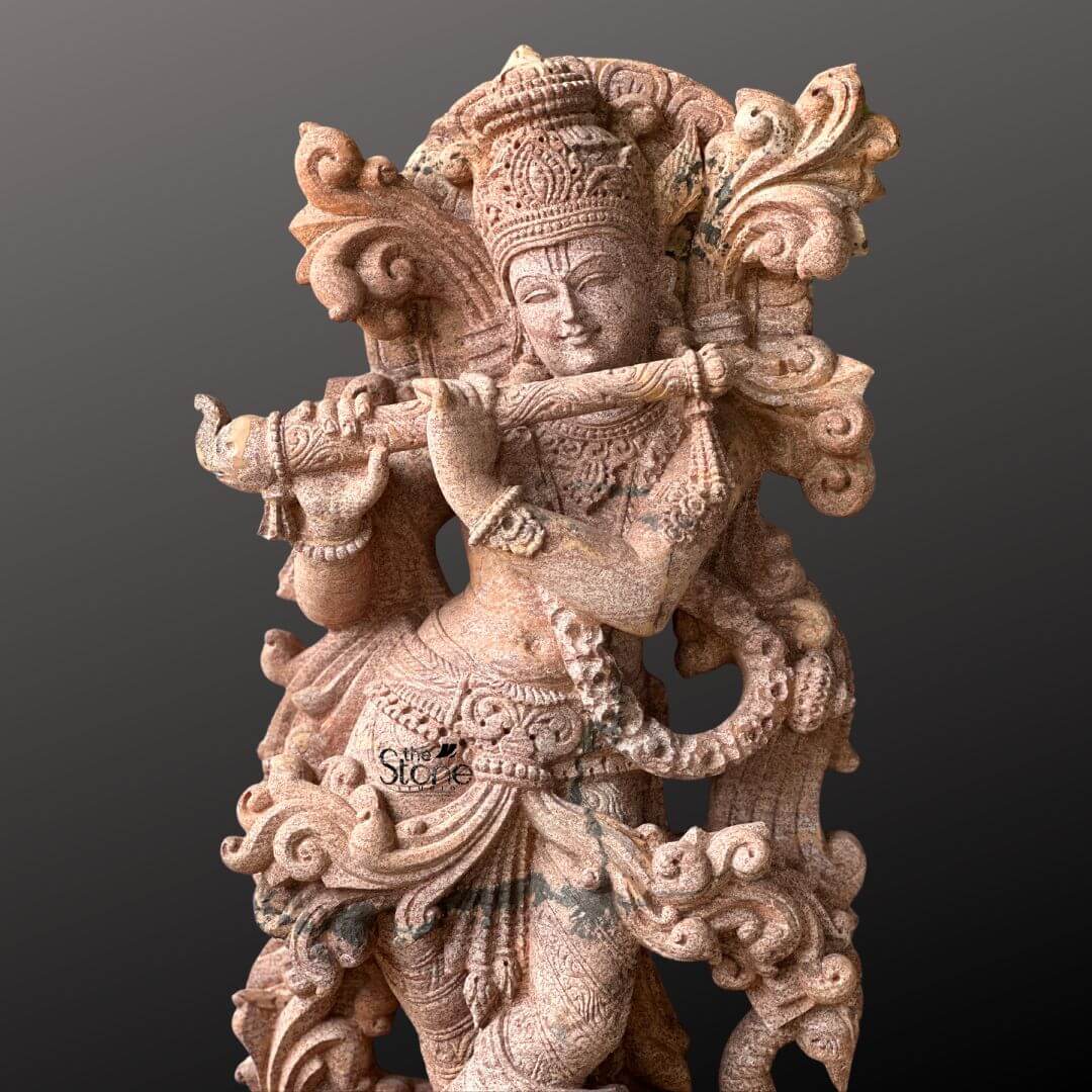 Standing Krishna Stone Statue with Flute 3.5ft 8 A close-up captures the upper portion of a magnificent 3.5-foot Standing Krishna Statue with Flute meticulously carved from sandstone presented against a deep contrasting background. Lord Krishna is depicted gracefully playing his bansuri with his head tilted slightly and a serene captivating expression on his face. His hands are delicately positioned to hold the flute highlighting his iconic role as the divine musician. The sandstone sculpture is a marvel of intricate detail especially evident in its rich ornamentation. Krishna wears an elaborate crown exquisitely carved with patterns and possibly featuring a peacock feather a quintessential symbol of his divine persona. His upper body is adorned with finely carved jewelry including multiple necklaces armlets and a decorative waist belt all rendered with precision in the warm-toned stone. The textured surface of the sandstone emphasizes the fine lines and curves of the drapery around his waist adding to the sculpture's tactile and visual appeal. An ornate swirling design forms a decorative frame behind his upper body possibly representing auspicious foliage or celestial elements. This detailed image of the 3.5-foot sandstone Standing Krishna Statue with Flute is perfectly suited for an article focusing on such devotional and artistic sculptures. The warm earthy tones of the sandstone beautifully capture the divine charm of Krishna making the flute-playing pose come alive. The close-up perspective allows viewers to appreciate the masterful carving from the subtle expression on Krishna's face to the intricate patterns on his crown and jewelry underscoring the statue's spiritual and aesthetic significance.