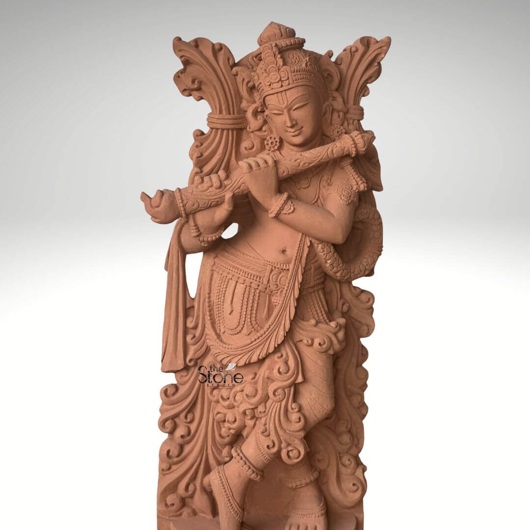 Lord Krishna Murti with Flute 3.5ft 6 A captivating 3.5-foot standing Krishna stone sculpture meticulously carved from sandstone depicted playing his flute as highlighted in an article about such sculptures. The entire statue rendered in the natural reddish-brown hue of sandstone exudes an earthy warm and timeless aesthetic. Lord Krishna is shown standing in his iconic Tribhanga pose with one leg crossed gracefully in front of the other an expression of divine playfulness and charm. He holds a flute to his lips with both hands appearing to play an enchanting melody. The carving details on Krishna's attire including his dhoti sash and intricate jewelry such as necklaces armlets and ankle bracelets are exceptionally refined. He wears an elaborate crown and a garland or Vanamala drapes diagonally across his body. The flowing drapery of his garments is skillfully sculpted adding a sense of movement and grace to the figure. Behind Krishna an intricately carved design possibly representing foliage or an artistic prabhavali further enhances the visual richness of the murti. The sculpture stands on a circular ornate pedestal also crafted from sandstone featuring traditional Indian architectural motifs and decorative patterns adding to its grandeur and stability. The texture of the sandstone is visible throughout providing depth and authenticity to the artwork. This 3.5-foot sandstone Krishna statue is a powerful representation of devotion and artistic mastery embodying the spiritual and aesthetic value discussed in the article making it an ideal centerpiece for spiritual veneration and artistic appreciation.