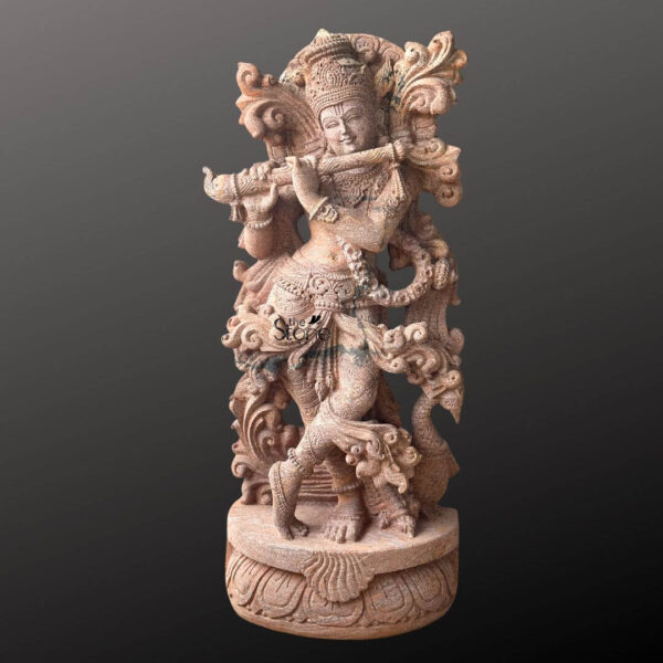 A beautifully carved 3.5-foot standing Krishna statue made from sandstone is showcased against a dark featureless background that emphasizes its intricate details and warm reddish-brown hue. Lord Krishna is depicted in his iconic Tribhanga pose with a gentle sway in his body playing his divine flute. His left leg is gracefully crossed in front of his right and his hands are raised to hold the instrument conveying a sense of enchanting melody and movement. The sandstone statue is replete with exquisite ornamentation reflecting the traditional imagery of Krishna. He wears an elaborate crown adorned with what appears to be a peacock feather a symbol strongly associated with the deity. Delicate jewelry including necklaces armlets waistbands and anklets is carved into the stone highlighting his divine beauty. The sculptor has paid meticulous attention to the flowing drapery of his dhoti and the ornate sash which seem to billow around him adding a dynamic quality to the static stone. Behind Krishna an intricately carved arch or halo possibly representing foliage or a peacock's fan frames his upper body enhancing his regal appearance. This 3.5-foot sandstone Krishna statue playing the flute, is an ideal subject for an article about standing Krishna stone statues. The warm tones of the sandstone contribute to its inviting and devotional appeal making it a perfect centerpiece for a temple home altar or garden. The detailed carving and the lifelike portrayal of Krishna in his most beloved form underscore the statue's artistic and spiritual significance truly bringing the essence of the divine flutist to life.