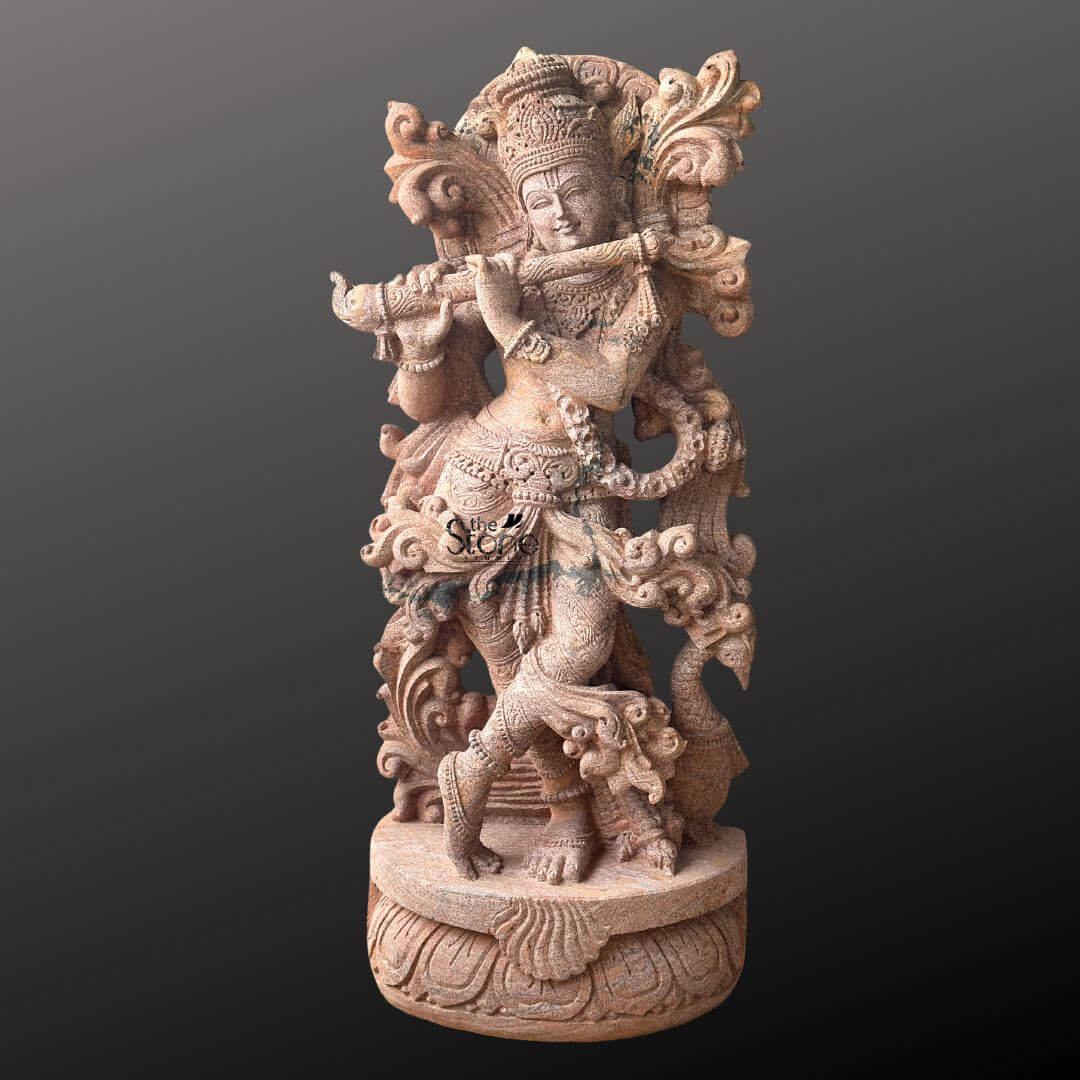 Standing Krishna Stone Statue with Flute 3.5ft 1 A beautifully carved 3.5-foot standing Krishna statue made from sandstone is showcased against a dark featureless background that emphasizes its intricate details and warm reddish-brown hue. Lord Krishna is depicted in his iconic Tribhanga pose with a gentle sway in his body playing his divine flute. His left leg is gracefully crossed in front of his right and his hands are raised to hold the instrument conveying a sense of enchanting melody and movement. The sandstone statue is replete with exquisite ornamentation reflecting the traditional imagery of Krishna. He wears an elaborate crown adorned with what appears to be a peacock feather a symbol strongly associated with the deity. Delicate jewelry including necklaces armlets waistbands and anklets is carved into the stone highlighting his divine beauty. The sculptor has paid meticulous attention to the flowing drapery of his dhoti and the ornate sash which seem to billow around him adding a dynamic quality to the static stone. Behind Krishna an intricately carved arch or halo possibly representing foliage or a peacock's fan frames his upper body enhancing his regal appearance. This 3.5-foot sandstone Krishna statue playing the flute, is an ideal subject for an article about standing Krishna stone statues. The warm tones of the sandstone contribute to its inviting and devotional appeal making it a perfect centerpiece for a temple home altar or garden. The detailed carving and the lifelike portrayal of Krishna in his most beloved form underscore the statue's artistic and spiritual significance truly bringing the essence of the divine flutist to life.