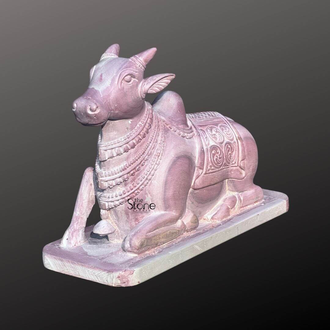 Stone Nandi Statue for Home Temple 8" 1 An exquisite 8-inch stone Nandi statue meticulously carved from pink stone presented against a dark background making it an ideal piece for a home temple as highlighted in the accompanying article. The entire sculpture rendered in a soft warm pinkish-brown hue of natural stone exudes a gentle and serene aesthetic. Nandi Lord Shiva's sacred bull is depicted in a traditional reclining posture symbolizing unwavering devotion patience and strength. The statue showcases remarkable detail in its carving. Nandi is adorned with a decorative saddle or blanket intricately carved with traditional Indian motifs and numerous strands of jewelry around his neck and head. His horns are neatly sculpted and his ears are gently laid back conveying a sense of calm and attentiveness. The eyes are subtly defined adding to the serene expression. The natural texture and subtle color variations of the pink stone are visible throughout adding authenticity and depth to the artwork. The Nandi statue rests on a simple rectangular base also carved from the same pink stone ensuring stability and a cohesive look. The smooth contours and fine details emphasize a deep sense of tranquility and spiritual harmony. This 8-inch pink stone Nandi statue is a powerful representation of devotion and artistic mastery perfectly suited for spiritual veneration in a home temple embodying the revered companion of Lord Shiva as discussed in the accompanying article.An exquisite 8-inch stone Nandi statue meticulously carved from pink stone presented against a dark background making it an ideal piece for a home temple as highlighted in the accompanying article. The entire sculpture rendered in a soft warm pinkish-brown hue of natural stone exudes a gentle and serene aesthetic. Nandi Lord Shiva's sacred bull is depicted in a traditional reclining posture symbolizing unwavering devotion patience and strength. The statue showcases remarkable detail in its carving. Nandi is adorned with a decorative saddle or blanket intricately carved with traditional Indian motifs and numerous strands of jewelry around his neck and head. His horns are neatly sculpted and his ears are gently laid back conveying a sense of calm and attentiveness. The eyes are subtly defined adding to the serene expression. The natural texture and subtle color variations of the pink stone are visible throughout adding authenticity and depth to the artwork. The Nandi statue rests on a simple rectangular base also carved from the same pink stone ensuring stability and a cohesive look. The smooth contours and fine details emphasize a deep sense of tranquility and spiritual harmony. This 8-inch pink stone Nandi statue is a powerful representation of devotion and artistic mastery perfectly suited for spiritual veneration in a home temple embodying the revered companion of Lord Shiva as discussed in the accompanying article.