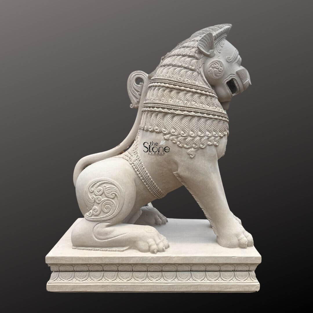 Traditional Stone Temple Lion Statue 7ft 8 A magnificent 7-foot Stone Simha Statue intricately carved from off-white sandstone is presented in a stately side profile against a dark plain background emphasizing its grandeur and intricate details perfectly suited for a temple entrance. The statue depicts a powerful lion in a seated alert posture embodying strength guardianship and regal authority making it an iconic and protective figure for sacred entrances. The light natural hue of the sandstone allows the elaborate carvings to stand out vividly. The lion's features are highly stylized and ornamental typical of traditional temple art. Its mane is intricately carved with layers of textured patterns resembling scales or braided hair and its powerful neck is adorned with decorative bands. The lion's face seen in profile features prominent eyes, a strong snout and an open mouth conveying a sense of watchful guardianship and a roaring presence. Its muscular body is embellished with swirling motifs and decorative elements on its shoulders and hindquarters. The tail curls elegantly along its back terminating in an ornamental flourish. The entire sculpture rests upon a robust, multi-tiered base also adorned with intricate patterns providing a solid foundation and enhancing its imposing presence at a temple entrance. This 7-foot off-white sandstone Stone Simha Statue is a profound representation of power, protection, and divine authority, making it an ideal guardian for a temple entrance. The exceptional craftsmanship in sandstone not only makes it a visually striking piece but also imbues it with deep cultural and religious significance. This exquisitely detailed statue serves as a commanding sentinel, radiating strength and an ancient spiritual presence, embodying centuries of artistic tradition and architectural grandeur within Hindu temples.