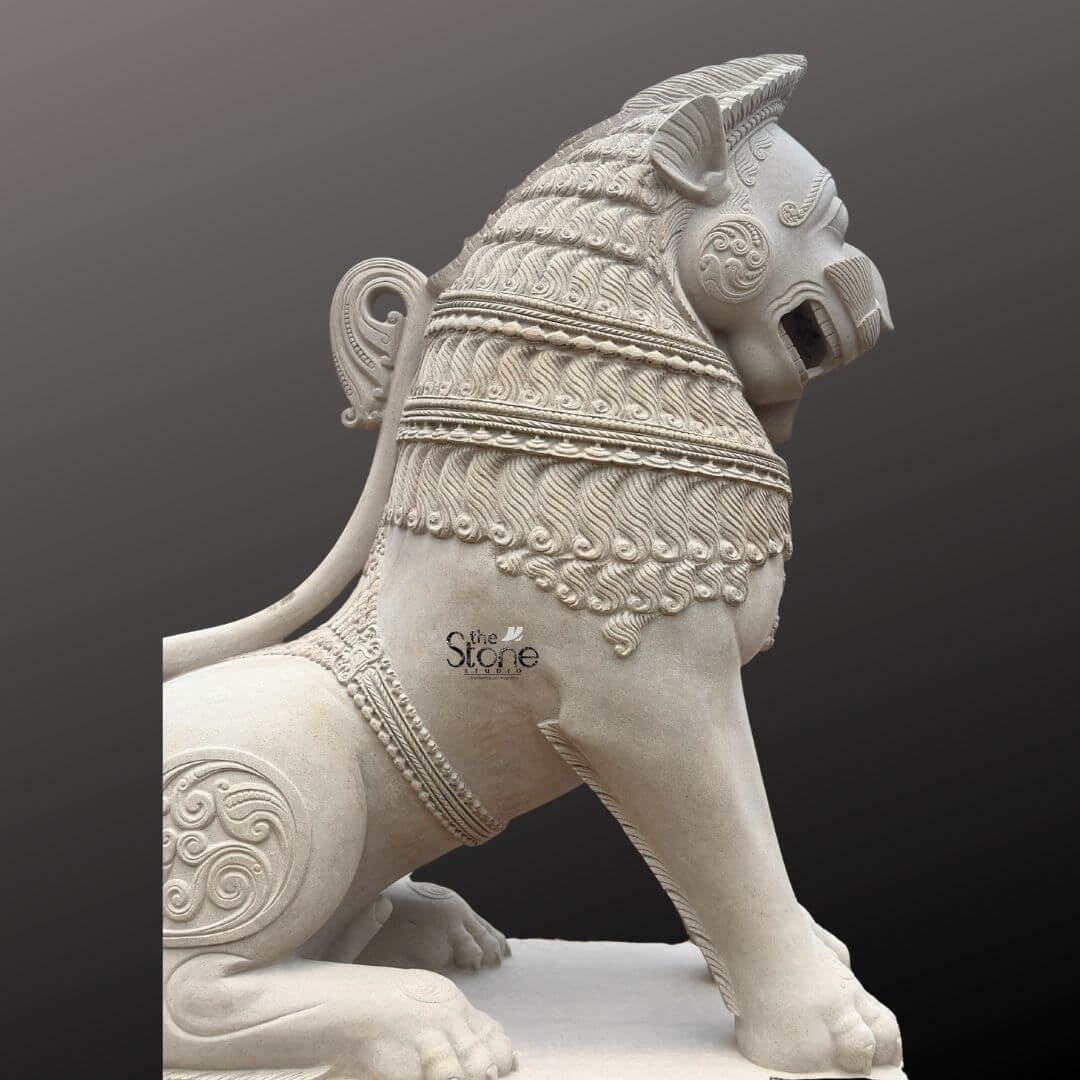 Traditional Stone Temple Lion Statue 7ft 10 A magnificent 7-foot Temple Guardian Lion Stone Statue intricately carved from off-white sandstone is presented in a stately side profile against a dark plain background emphasizing its grandeur and commanding presence. The statue depicts a powerful lion in a seated alert posture embodying strength guardianshi, and regal authority, making it an iconic and protective figure for sacred entrances. The light, natural hue of the sandstone allows the elaborate carvings to stand out vividly. The lion's features are highly stylized and ornamental typical of traditional temple art. Its mane is intricately carved with layers of textured patterns resembling scales or braided hair and its powerful neck is adorned with decorative bands. The lion's face, seen in profile, features prominent eyes, a strong snout, and an open mouth conveying a sense of watchful guardianship and a roaring presence. Its muscular body is embellished with swirling motifs and decorative elements on its shoulders and hindquarters. The tail curls elegantly along its back terminating in an ornamental flourish. The entire sculpture rests upon a robust multi-tiered base providing a solid foundation and enhancing its imposing presence as a temple guardian. This 7-foot off-white sandstone Temple Guardian Lion Stone Statue is a profound representation of power, protection, and divine authority, making it an ideal guardian for a temple. The exceptional craftsmanship in sandstone not only makes it a visually striking piece but also imbues it with deep cultural and religious significance. This exquisitely detailed statue serves as a commanding sentinel, radiating strength and an ancient spiritual presence, embodying centuries of artistic tradition and architectural grandeur within Hindu temples.