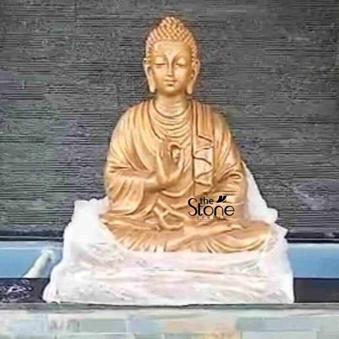 Metal Buddha 3ft 6 A radiant golden Buddha statue crafted for outdoor display is prominently featured in what appears to be a terrace garden setting designed to bring peace and serenity to an urban environment like Bangalore. The statue depicts the Buddha seated in a classic meditative posture with his right hand in the Vitarka Mudra symbolizing teaching and intellectual discussion while his left hand rests in his lap palm upwards signifying meditation. The lustrous golden finish of the statue stands out vibrantly against a dark textured tile wall creating a striking visual contrast. The Buddha's face is carved with an expression of profound serenity and wisdom with downcast eyes suggesting deep contemplation. His ushnisha and tightly curled hair are meticulously detailed indicative of a finely crafted piece. The flowing drapery of his robes is rendered with soft natural folds adding to the realism and grace of the figure. The statue rests on a simple white fabric or base which in turn sits on a dark possibly stone platform providing a dignified foundation. The horizontal blue line behind the statue might indicate a pool or water feature enhancing the tranquil ambiance often desired in terrace gardens. This golden Buddha statue is an exceptional piece that brings an immediate sense of calm and spiritual harmony to any outdoor space particularly suitable for terrace gardens in Bangalore. Its golden finish radiates positive energy and makes it a striking focal point while its durable construction ensures longevity in outdoor conditions. As a symbol of enlightenment and peace this Buddha statue is not only an artistic masterpiece but also a significant spiritual icon inviting contemplation and fostering a tranquil atmosphere for relaxation and spiritual connection.