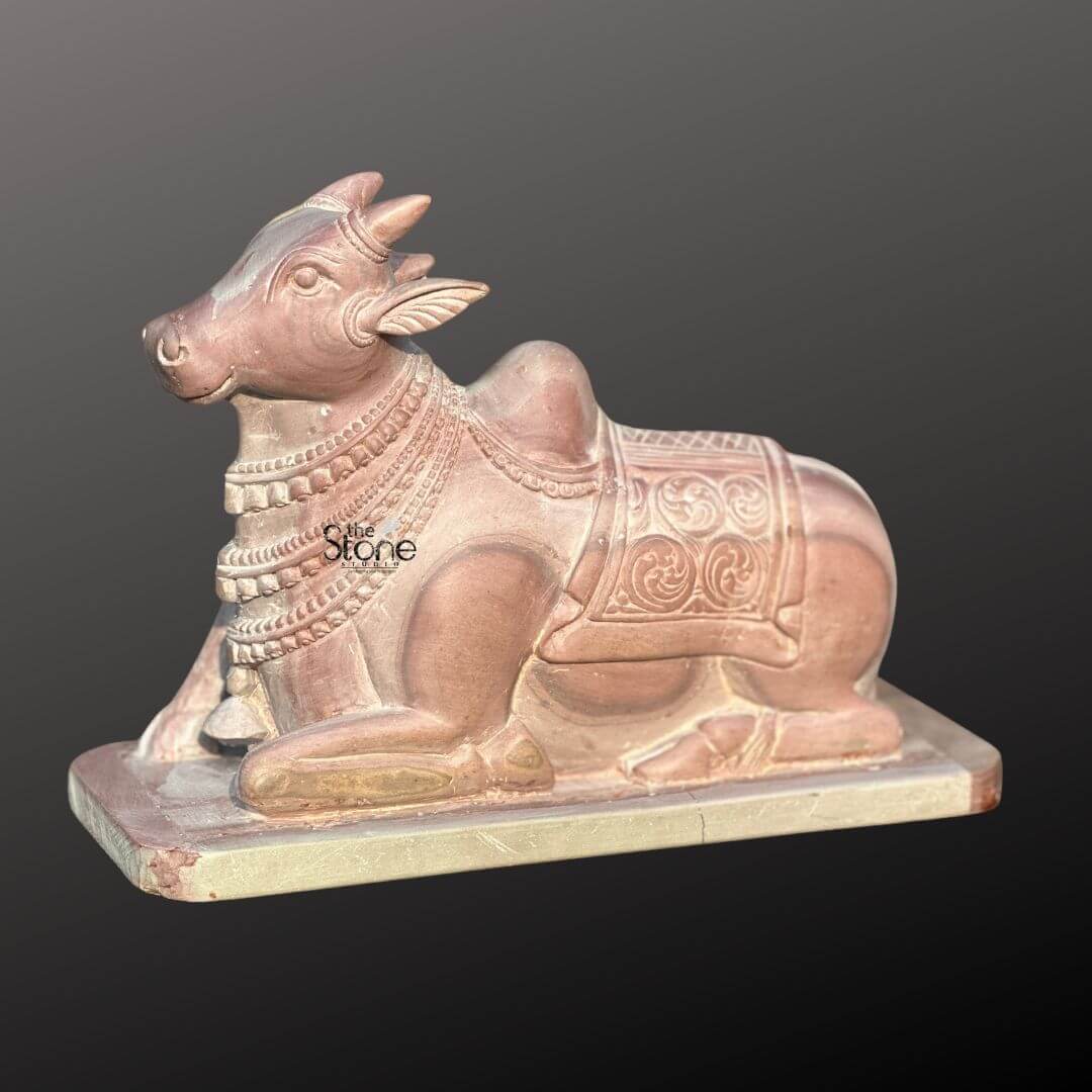 Stone Nandi Statue for Home Temple 8" 8 A meticulously carved 8-inch traditional Nandi bull idol crafted from natural pink stone is presented in a serene side profile against a dark contrasting background. The statue depicts Nandi in a classic reclining pose with its head gracefully turned to the lef emanating an aura of peace and devotion. The inherent pink and reddish tones of the stone are beautifully highlighted showcasing the subtle striations and natural variations that lend uniqueness and character to this sacred sculpture. The Nandi idol is richly adorned with intricate traditional embellishments including multiple strands of beaded necklaces draped around its neck and a detailed ceremonial blanket over its back. The blanket features elaborate scrollwork and geometric patterns meticulously carved to add depth and authenticity to the figure. Its distinct humped back a hallmark of the revered Indian bull is clearly visible and the overall form is sculpted with a gentle elegance that reflects its divine status. This 8-inch traditional Nandi bull idol in pink stone serves as a powerful symbol of loyalty strength and unwavering devotion in Hindu tradition. Its compact size makes it a versatile piece perfect for a home mandir meditation space or as a cultural art display. The skillful craftsmanship combined with the natural beauty of the pink stone makes this Nandi statue not just a decorative item but a meaningful artifact that resonates with spiritual significance and ancient cultural heritage.