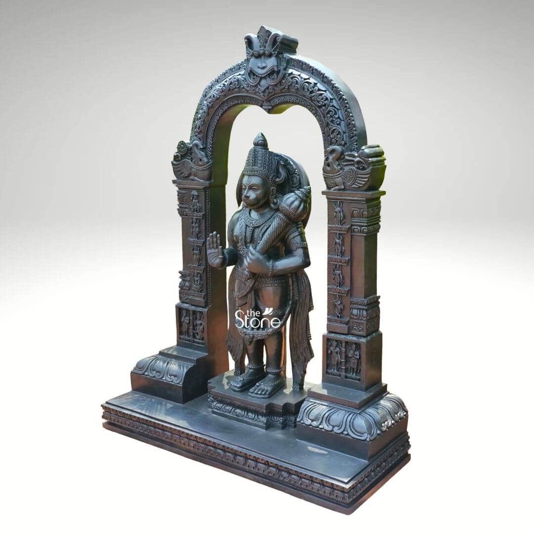 Standing Bajrangbali Statue 4ft 8 An impressive black stone Traditional Standing Hanuman Idol powerfully holding his Gada and framed by an intricately carved temple arch is presented against a plain light background. This stunning black stone statue captures Lord Hanuman in a dynamic standing posture exuding an aura of immense strength unwavering devotion and courage. The dark polished surface of the stone provides a dramatic contrast enhancing the intricate details of the sculpture and its elaborate architectural frame making it a profound focal point for worship and admiration. Lord Hanuman is depicted with his characteristic features including a majestic crown prominent ear ornaments and a strong muscular physique that symbolizes his extraordinary power. His right hand is raised in the Abhaya Mudra offering blessings and protection to devotees while his left hand firmly holds his Gada resting on his shoulder symbolizing his immense power and readiness to vanquish evil. A garland of beads adorns his neck and a decorative sash drapes across his body all carved with meticulous precision. He stands on a lotus pedestal which is integrated into a multi-tiered base adorned with smaller figures and decorative carvings all contributing to the grandeur of the piece. The intricately carved temple archway above him is equally detailed featuring divine figures floral motifs and a Kirtimukha at the apex flanked by mythical creatures. This black stone Traditional Standing Hanuman Idol with Gada and its ornate carved arch is a profound representation of Lord Hanuman the epitome of strength devotion and selfless service. The exceptional craftsmanship in black stone not only makes it a visually striking piece but also embodies deep cultural and religious significance offering a powerful focal point for devotion and prayer. The rich detailing of the deity and his surrounding archway reflects centuries of tradition making it a truly remarkable and revered artifact that brings blessings protection and a spiritual presence into any sacred space.