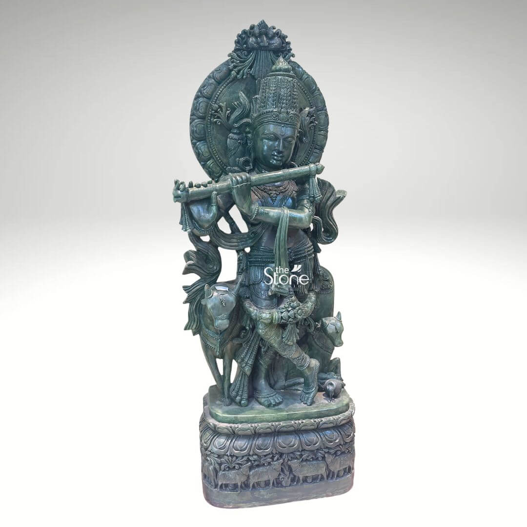 Green Stone Krishna with Cow Statue 4ft 7 A magnificent Venugopal Krishna Green Stone Statue meticulously carved from greenstone stands prominently against a clean white background showcasing exquisite detail and artistry. Lord Krishna in his form as Venugopal is depicted in his iconic Tribhanga pose gracefully playing his flute his body angled with a serene expression. Beside him a cow and a calf are intricately carved reflecting his identity as a divine cowherd an essential aspect of the Venugopal Krishna theme. The greenstone material gives the entire sculpture a distinctive earthy and revered appearance. The greenstone Venugopal Krishna statue is rich with traditional ornamentation. Krishna wears an elaborate crown likely adorned with a peacock feather a symbol strongly associated with him. His form is embellished with finely carved jewelry including necklaces armlets, waistbands and anklets. The flowing drapery of his dhoti and sashes adds to the dynamic quality of the pose. Behind Krishna a circular aureole or halo frames his head often symbolizing divinity and a larger ornate arch rises above featuring intricate patterns. The cow and calf are rendered with gentle expressions symbolizing prosperity devotion and the tender bond between mother and offspring and they are surrounded by carved foliage and other natural elements. This Greenstone Venugopal Krishna Green Stone Statue is an ideal visual for an article detailing such divine sculptures. The statue rests on a multi-tiered intricately carved base which features a band of smaller animal figures possibly more cows further enhancing the pastoral theme. The harmonious composition of Krishna the cow and calf and the detailed base in greenstone creates a powerful and spiritually uplifting piece embodying devotion divine love and the sacred bond between Krishna animals and nature perfect for a temple or a home seeking a touch of spiritual grace associated with Venugopal Krishna.