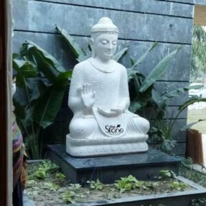 A magnificent 6-foot White Stone Buddha Statue specifically carved from white sandstone is majestically positioned outdoors against a modern textured grey stone wall likely in a garden or courtyard setting in Delhi. The Buddha is depicted in a serene seated posture embodying deep meditation and tranquility. His right hand is raised in the Abhaya Mudra, a gesture of fearlessness and reassurance while his left hand rests gracefully in his lap palms open. The white sandstone Buddha statue exudes purity and calm with meticulously carved details. The Buddha's face features downcast eyes a gentle smile and elongated earlobes conveying profound inner peace and wisdom. His hair is intricately sculpted in tight curls culminating in the ushnisha the cranial bump symbolizing enlightenment. The flowing robes are rendered with delicate pleats and folds creating a sense of natural drapery that adds to the statue's lifelike quality. The statue sits on a simple yet elegant base adorned with subtle lotus petal motifs further enhancing its spiritual aesthetic. This impressive 6-foot white sandstone Buddha statue is a perfect example for an article discussing such statues in Delhi. The pristine white stone contrasts beautifully with the darker background wall and the surrounding green foliage making it a prominent and calming focal point. The statue's substantial size and serene presence make it an ideal choice for a home garden or public space inviting contemplation and bringing a sense of peace and spiritual harmony to its environment.