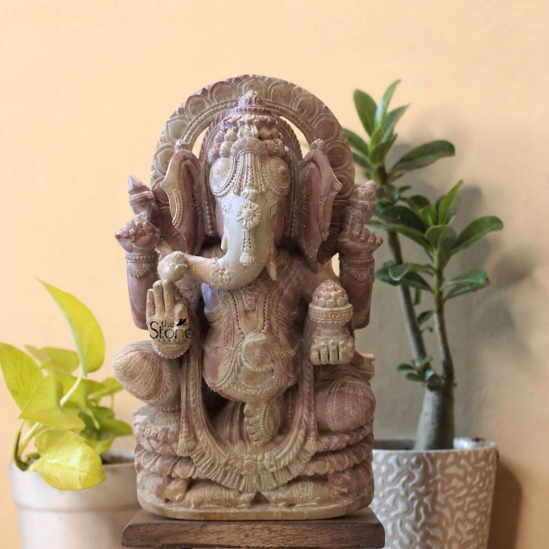 Pink Stone Ganpati Idol for Home 12" 3 A finely detailed twelve-inch tall Ganesha statue hand-carved from natural pink stone is the central focus of this studio photograph. The statue depicts the elephant-headed Hindu deity in a seated meditative pose with his left leg folded and right leg pendent resting upon a multi-tiered lotus-carved pedestal which is itself mounted on a simple dark wooden block. The pink stone displays natural variations in hue ranging from soft rose to deeper mauve giving the idol a warm and earthy texture. Ganesha is richly adorned with intricate deeply carved traditional jewelry including elaborate necklaces armbands anklets and a richly patterned crown that frames his head reflecting the skill of the artisan. The four-armed deity holds symbolic attributes in his hands all rendered with meticulous precision. His upper right hand holds a goad while his upper left hand holds a noose. His lower right hand is raised in the Abhaya mudra a gesture of blessing and protection and his lower left hand holds a bowl overflowing with modaks. Ganesha's trunk curves gently to his left sampling the sweets in his palm. The carving extends to his garments which are textured to resemble rich pleated fabric held by a jeweled girdle. Behind his head is a decorative prabhavali with floral and geometric carvings, enhancing the divine aura of the figure. This twelve-inch pink stone Ganesha statue is an ideal size for a home altar a prominent shelf or as a decorative centerpiece bringing a sense of spiritual tranquility and cultural artistry to any interior space. The warm pink color of the stone offers a gentle and inviting aesthetic making it a beautiful addition to diverse home decor styles. The detailed craftsmanship and traditional iconography make this Ganesha idol a meaningful choice for devotees and collectors alike representing wisdom prosperity and the removal of obstacles. The statue's durable stone construction ensures its longevity as a cherished spiritual and decorative piece.