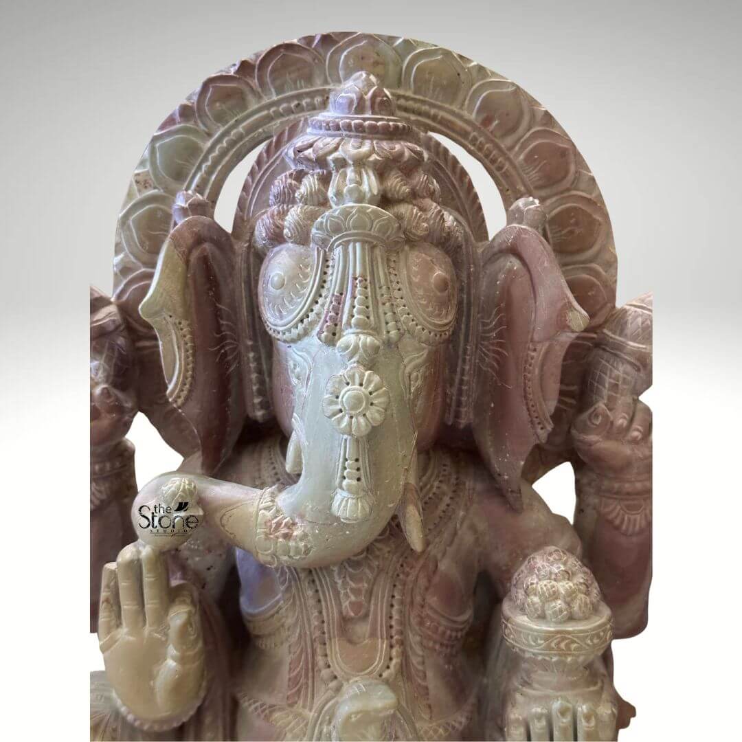 Pink Stone Ganpati Idol for Home 12" 5 A detailed close-up photograph captures a hand-carved stone Ganesh Murti measuring twelve inches in height. The statue is sculpted from a natural pink stone which exhibits a warm variegated hue ranging from soft rose to deeper mauve with subtle crystalline inclusions visible on the surface. The elephant-headed deity is depicted in a seated meditative pose with his trunk gently curving to his left holding a sweet modak. His eyes are partially closed in contemplation and he wears an elaborate intricately carved crown with floral and geometric motifs that reflect the artisan's skill. The stone surface is smooth emphasizing the sculptural details. The four-armed figure is richly adorned with traditional ornaments all rendered in deep relief carving. He wears multiple layered necklaces large circular earrings armlets and bracelets. His garments are textured to resemble draped fabric held by a jeweled girdle. In his upper right hand he holds an axe while his upper left hand holds a noose. His lower right hand is raised in the Abhaya mudra a gesture of blessing and protection and his lower left hand holds a bowl overflowing with modaks which his trunk is sampling. Behind his head, a decorative prabhavali frames his form adorned with intricate lotus petal carvings. This twelve-inch pink stone Ganesha statue is an exemplary piece of Indian stone carving art making it a perfect subject for an article on Stone Ganesh Murtis. The compact size indicates its suitability for a home altar a prominent shelf or as a focal point in a smaller room bringing a sense of spiritual tranquility and aesthetic beauty to any space. The choice of pink stone adds a gentle inviting color to the decor. The detailed iconography and traditional craftsmanship showcased in this image highlight the enduring cultural and religious significance of Ganesha in Indian households. The photograph's clear focus on the statue's texture and form provides a comprehensive view for the viewer.
