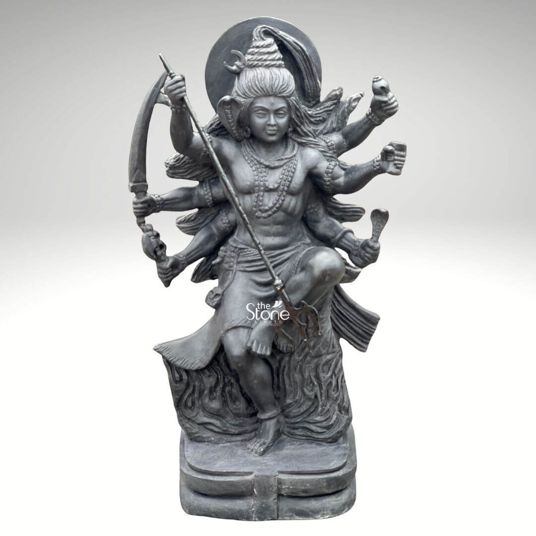 2 Feet Tandava Shiva Statue 8 A full-body studio photograph of a magnificent 2-foot tall Black Stone Tandava Shiva Statue masterfully hand-carved from a single block of dense polished black stone. The statue captures Lord Shiva in the dynamic and powerful Ananda Tandava pose the dance of bliss set upon a multi-tiered lotus pedestal. Shiva is depicted with ten arms fanned out to convey movement holding various divine attributes including a trident and damaru drum. His matted hair is piled high adorned with a crescent moon and a serpent and flares out dramatically to emphasize the energy of the dance. The intricate details of the carving from the folds of his garment to the expressions on his many faces, are sharply defined, showcasing the exquisite craftsmanship. The black stone used for the statue has a deep rich hue with a subtle waxy luster giving it a powerful and reverent presence. The ten arms are arranged with precision some raised in gestures of protection and blessing others holding attributes like a conch shell a fire pot and a scripture. Shiva's right leg is raised bent at the knee while his left leg is planted firmly on the demon Muyalakan who lies subdued beneath his foot. The dynamism of the dance is conveyed through the arching of his body and the intricate arrangement of his limbs creating a sense of controlled power and divine ecstasy. This 2-foot black stone Dancing Shiva statue stands as a testament to Indian temple art and a perfect centerpiece for any sacred space. Its compact size allows for placement on an altar or a display table where its intricate details can be admired. The dark monolithic stone gives it a timeless quality while the powerful iconography of the Tandava pose evokes a sense of cosmic energy and spiritual liberation. The photograph presents the statue against a clean neutral white background ensuring the focus remains entirely on the form texture and spiritual significance of the black stone sculpture.