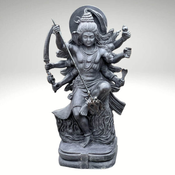 A full-body studio photograph of a magnificent 2-foot tall Tandava Shiva Statue masterfully hand-carved from a single block of dense polished black stone. The statue captures Lord Shiva in the dynamic and powerful Ananda Tandava pose the dance of bliss set upon a multi-tiered lotus pedestal. Shiva is depicted with ten arms fanned out to convey movement holding various divine attributes including a trident and damaru drum. His matted hair is piled high, adorned with a crescent moon and a serpent and flares out dramatically to emphasize the energy of the dance. The intricate details of the carving from the folds of his garment to the expressions on his many faces are sharply defined showcasing the exquisite craftsmanship. The black stone used for the statue has a deep rich hue with a subtle waxy luster giving it a powerful and reverent presence. The ten arms are arranged with precision some raised in gestures of protection and blessing others holding attributes like a conch shell a fire pot, and a scripture. Shiva's right leg is raised, bent at the knee, while his left leg is planted firmly on the demon Muyalakan who lies subdued beneath his foot. The dynamism of the dance is conveyed through the arching of his body and the intricate arrangement of his limbs creating a sense of controlled power and divine ecstasy. This 2-foot black stone Tandava Shiva statue stands as a testament to Indian temple art and a perfect centerpiece for any sacred space. Its compact size allows for placement on an altar or a display table where its intricate details can be admired. The dark monolithic stone gives it a timeless quality while the powerful iconography of the Tandava pose evokes a sense of cosmic energy and spiritual liberation. The photograph presents the statue against a clean neutral white background ensuring the focus remains entirely on the form texture and spiritual significance of the black stone sculpture.