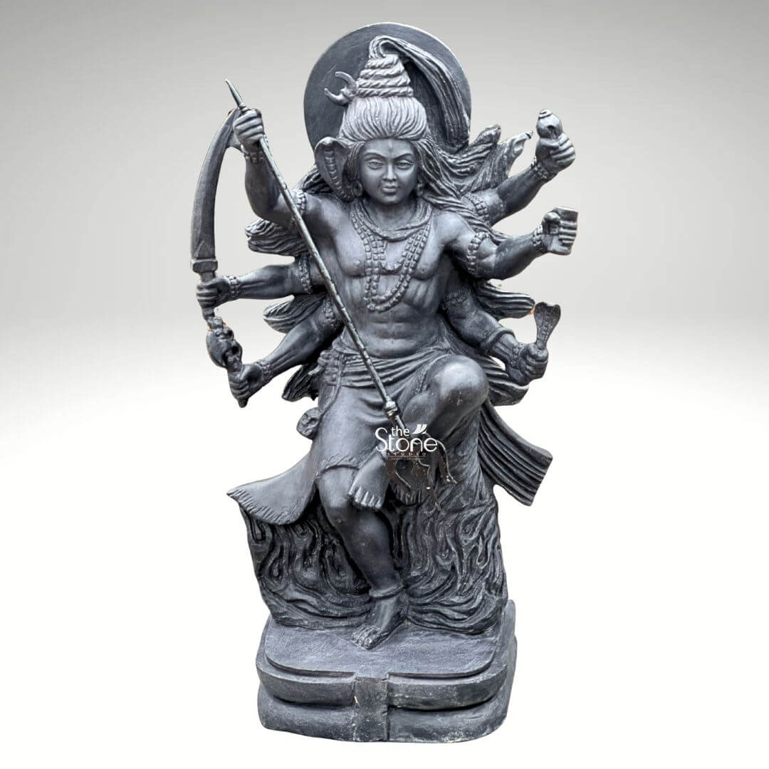 2 Feet Tandava Shiva Statue 1 A full-body studio photograph of a magnificent 2-foot tall Tandava Shiva Statue masterfully hand-carved from a single block of dense polished black stone. The statue captures Lord Shiva in the dynamic and powerful Ananda Tandava pose the dance of bliss set upon a multi-tiered lotus pedestal. Shiva is depicted with ten arms fanned out to convey movement holding various divine attributes including a trident and damaru drum. His matted hair is piled high, adorned with a crescent moon and a serpent and flares out dramatically to emphasize the energy of the dance. The intricate details of the carving from the folds of his garment to the expressions on his many faces are sharply defined showcasing the exquisite craftsmanship. The black stone used for the statue has a deep rich hue with a subtle waxy luster giving it a powerful and reverent presence. The ten arms are arranged with precision some raised in gestures of protection and blessing others holding attributes like a conch shell a fire pot, and a scripture. Shiva's right leg is raised, bent at the knee, while his left leg is planted firmly on the demon Muyalakan who lies subdued beneath his foot. The dynamism of the dance is conveyed through the arching of his body and the intricate arrangement of his limbs creating a sense of controlled power and divine ecstasy. This 2-foot black stone Tandava Shiva statue stands as a testament to Indian temple art and a perfect centerpiece for any sacred space. Its compact size allows for placement on an altar or a display table where its intricate details can be admired. The dark monolithic stone gives it a timeless quality while the powerful iconography of the Tandava pose evokes a sense of cosmic energy and spiritual liberation. The photograph presents the statue against a clean neutral white background ensuring the focus remains entirely on the form texture and spiritual significance of the black stone sculpture.