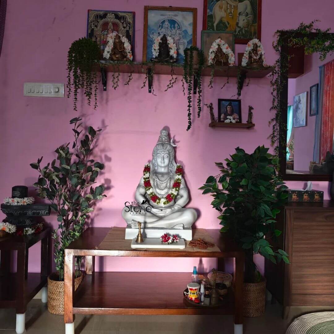 A reverent eye-level photograph captures a beautifully decorated home shrine dedicated to Lord Shiva centered around a 2 Feet Fibre Shiva Idol. The idol hand-painted in a matte white finish with subtle grey shading to define the musculature and traditional ornamentation depicts Shiva in a serene meditative padmasana pose. He is adorned with a vibrant fresh garland of red and white flowers contrasting with the white material. A miniature brass cobra hood is integrated above his head and the sacred river Ganges flows from his matted hair. The idol rests on a simple yet elegant wooden altar table positioned in front of a warm pink-painted wall creating a peaceful focal point for worship. The Fibre Shiva Idol is a substantial piece measuring approximately 2 feet in height making it perfectly scaled for a home puja room or personal altar space without being overpowering. The use of fiberglass construction ensures the statue is lightweight yet durable allowing for easier handling and placement compared to a traditional stone sculpture of similar size while still retaining a high level of detailed craftsmanship in the facial features and jewelry. The idol is flanked by two large realistic artificial potted plants housed in woven baskets adding a touch of nature and symmetry to the sacred space. Above the altar a wall-mounted wooden shelf displays various framed Hindu deities and spiritual images including depictions of Lakshmi Vishnu and a larger colorful painting of Shiva and Parvati on Nandi all adorned with strings of artificial green vines. The lower shelf of the altar table holds essential puja items including a small brass oil lamp prayer beads and various ritual offerings, emphasizing the idol's functional role in daily worship. The entire composition highlights the significance of this 2-foot fibre Shiva idol as an accessible and spiritually meaningful addition to a home blending traditional devotion with modern materials.