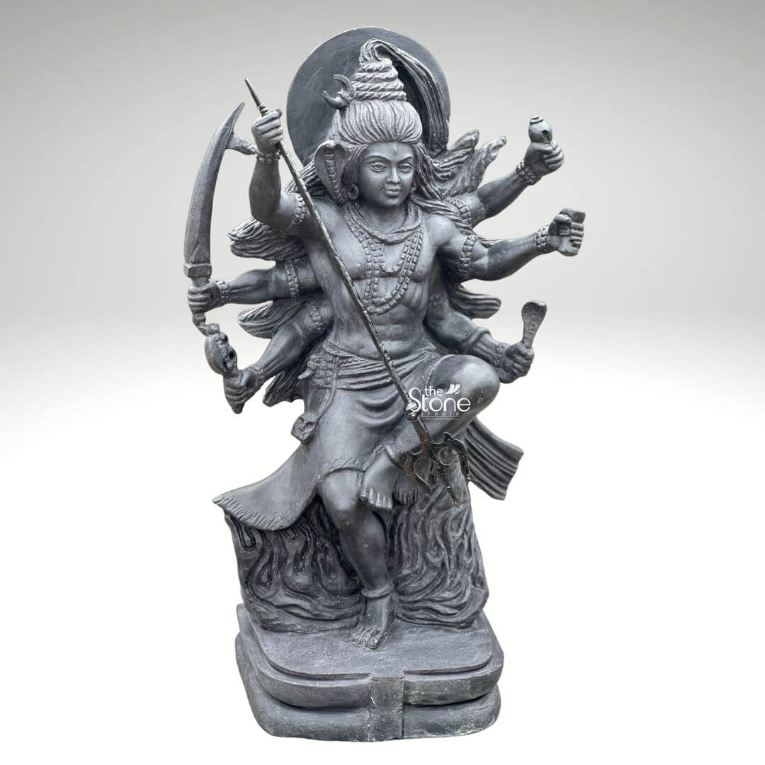 2 Feet Tandava Shiva Statue 2 A full-body studio photograph of a magnificent 2-foot tall Tandava Shiva Stone Statue masterfully hand-carved from a single block of dense polished black stone. The statue captures Lord Shiva in the dynamic and powerful Ananda Tandava pose the dance of bliss set upon a multi-tiered lotus pedestal. Shiva is depicted with ten arms fanned out to convey movement holding various divine attributes including a trident and damaru drum. His matted hair is piled high adorned with a crescent moon and a serpent and flares out dramatically to emphasize the energy of the dance. The intricate details of the carving from the folds of his garment to the expressions on his many faces are sharply defined showcasing the exquisite craftsmanship. The black stone used for the statue has a deep rich hue with a subtle waxy luster giving it a powerful and reverent presence. The ten arms are arranged with precision some raised in gestures of protection and blessing, others holding attributes like a conch shell a fire pot and a scripture. Shiva's right leg is raised bent at the knee while his left leg is planted firmly on the demon Muyalakan who lies subdued beneath his foot. The dynamism of the dance is conveyed through the arching of his body and the intricate arrangement of his limbs creating a sense of controlled power and divine ecstasy. This 2-foot black stone Tandava Shiva statue stands as a testament to Indian temple art and a perfect centerpiece for any sacred space. Its compact size allows for placement on an altar or a display table where its intricate details can be admired. The dark monolithic stone gives it a timeless quality while the powerful iconography of the Tandava pose evokes a sense of cosmic energy and spiritual liberation. The photograph presents the statue against a clean neutral white background ensuring the focus remains entirely on the form texture and spiritual significance of the black stone sculpture.