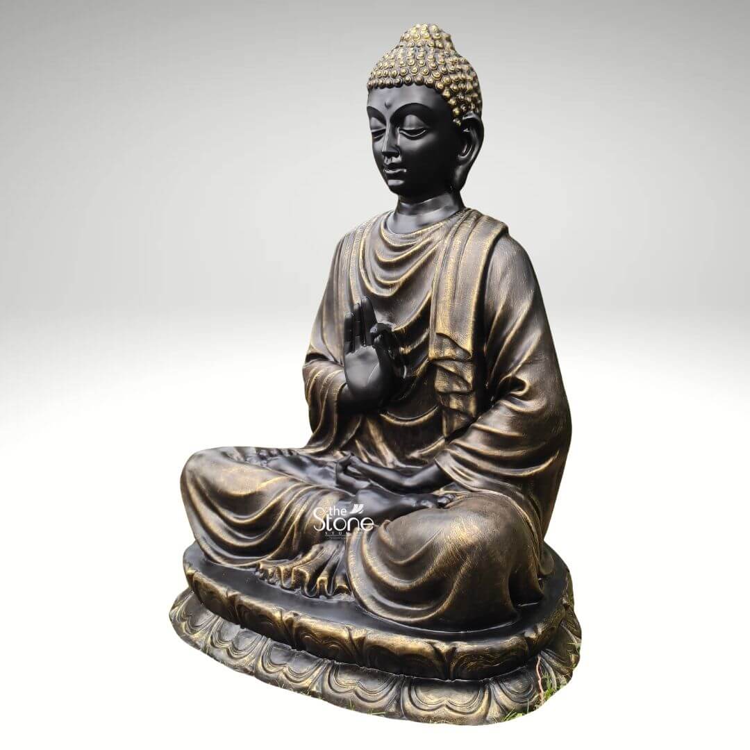 Large Fibre Buddha Statue 5ft 2 A 5 feet Buddha fibre statue in Vitarka Mudra is shown from a three-quarter side angle against a plain light grey background allowing the sculpture’s form and detailing to stand out clearly. The Buddha is seated cross-legged on a broad lotus pedestal with the right hand raised near the chest in Vitarka Mudra a gesture associated with teaching discussion and the transmission of wisdom. The left hand rests gently in the lap reinforcing the calm meditative posture. The face has a serene expression with softly closed eyes elongated earlobes and smooth balanced features that convey peace mindfulness and spiritual grace. The large fibre Buddha statue is finished in a rich combination of black and antique gold tones giving it a refined and dignified appearance. The curled hair is highlighted in gold creating contrast with the darker face and drawing attention to the head and upper features. Flowing robe folds drape naturally over the shoulders arms and legs with metallic gold accents enhancing the contours and craftsmanship of the sculpture. The lotus base beneath the seated figure is carved with repeating petal motifs adding both decorative elegance and symbolic meaning to the overall design. The angled view helps reveal the depth of the sculpture including the curve of the seated posture the layered drapery and the dimensional quality of the hand gesture. This image connects strongly to an article about a 5 Feet Buddha Statue in Vitarka Mudra by clearly showing the defining mudra large size and decorative appeal of the piece. The statue appears ideal for meditation rooms living spaces entryways, spiritual corners wellness studios and peaceful commercial interiors where a large Buddha statue can create a calming focal point. With its antique finish lotus pedestal and teaching gesture this 5 feet Buddha fibre statue combines spiritual symbolism elegant craftsmanship and timeless décor value making it a beautiful choice for those seeking a meaningful Buddha statue for home or meditation décor.