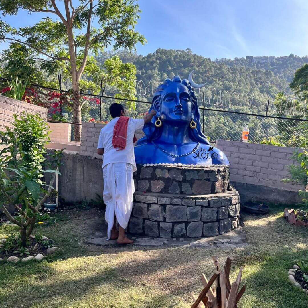 Blue Adiyogi Shiva Statue 4ft 6 A 4 feet Adiyogi Fibre Sculpture in Solan is installed as a striking spiritual focal point in an open garden setting surrounded by nature and hills. The sculpture depicts the iconic bust form of Adiyogi Lord Shiva finished in a vivid deep blue colour with calm facial features half-closed meditative eyes long flowing hair large earrings a crescent moon on the head and a beaded necklace across the chest. The serene expression and powerful scale of the fibre statue create a strong sense of devotion meditation and divine presence. The Adiyogi statue is placed on a circular stone pedestal made with dark natural-looking blocks giving the installation a grounded and temple-like appearance. A person dressed in traditional white clothing is standing near the statue appearing to perform a ritual or cleaning activity which adds a devotional and lived-in feeling to the scene. The garden around the sculpture includes trimmed grass small plants flowering shrubs stone borders and trees while the background opens into a scenic mountain landscape under a clear blue sky. This image connects beautifully to an article about an Adiyogi Fibre Sculpture in Solan by showing how a 4 feet fibre Adiyogi statue can enhance a garden meditation space resort, spiritual retreat farmhouse or outdoor landscape. The combination of the blue Shiva sculpture stone base natural greenery and hilly Solan backdrop highlights the statue’s suitability for peaceful outdoor décor and devotional installations. The overall scene conveys spirituality strength calmness and a deep connection between Lord Shiva, nature and meditative living.