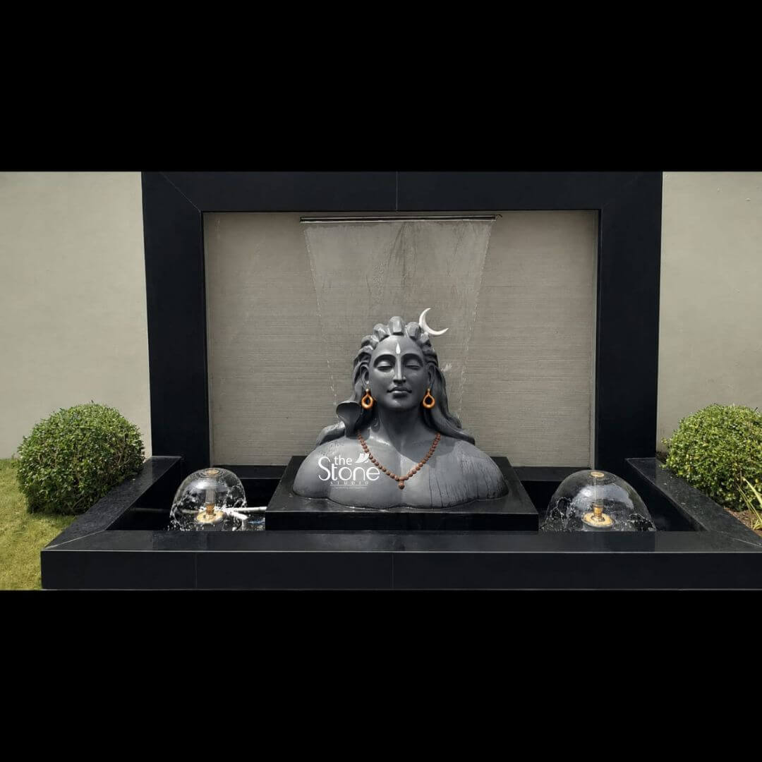 3 feet Adiyogi Statue 7 A striking three-foot Adiyogi bust statue is the divine centerpiece of a contemporary outdoor water fountain presented against a plain grey wall flanked by two manicured spherical shrubs. The statue crafted from durable dark grey fibre with a matte finish depicts the iconic form of Lord Shiva in his Adiyogi aspect. His face is serene and contemplative with downcast eyes a distinct third eye carved on his forehead and his matted hair intricately styled holding a crescent moon and adorned with a simple traditional brown bead necklace resting on his chest. The dark hue of the statue provides a powerful contrast to the surrounding elements. The Adiyogi statue is positioned on a raised monolithic black granite plinth which sits within a larger rectangular black granite basin. This basin forms the main reservoir of the water fountain featuring two symmetrical bubbling spherical water features on either side of the statue adding dynamic movement to the scene. A sheer sheet of water gracefully trickles down a textured grey granite back panel directly behind the statue creating a soothing curtain of sound and adding a layer of reflection on the water's surface. The entire structure is framed by a clean black geometric border enhancing its modern artistic appearance. This three-foot Fibre Adiyogi statue is an ideal subject for an article discussing Adiyogi Statue for Water Fountain perfectly demonstrating how such a sacred and iconic form can be integrated into a modern garden or patio design. The combination of the spiritual presence of Adiyogi with the calming element of cascading water creates a peaceful and meditative atmosphere. The durable fibre material ensures its suitability for outdoor elements as a long-lasting decorative accent while its detailed craftsmanship makes it a beautiful and meaningful focal point for any landscape. The composition evokes a sense of ancient tradition blended with contemporary style.