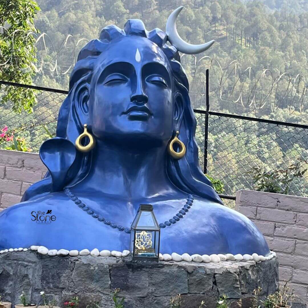 Blue Adiyogi Shiva Statue 4ft 9 A 4 feet Adiyogi fibre statue is shown as the central focus of an outdoor installation in Solan presented in a close-up view that highlights its calm and majestic presence. The sculpture depicts Adiyogi Lord Shiva in the iconic bust form with a serene meditative expression softly closed eyes and well-defined facial features that convey peace stillness and spiritual depth. The statue is finished in a striking deep blue tone with a white crescent moon placed on the head a sacred tilak mark on the forehead large circular earrings and a beaded necklace sculpted across the chest all of which add symbolic and devotional significance to the Adiyogi design. The 4 feet fibre Adiyogi statue is installed on a circular stone-clad pedestal made of dark natural-looking blocks with a border of smooth white pebbles arranged neatly along the top edge. At the front of the pedestal sits a small lantern-style decorative piece adding a subtle spiritual accent to the display. Behind the sculpture a wire fence and low masonry wall frame the space while the background opens to a scenic hillside covered with trees giving the setting a peaceful mountain atmosphere. The natural outdoor light enhances the rich blue finish of the statue and makes the facial contours and sculptural details stand out clearly. This image connects strongly to an article about an Adiyogi Statue in Solan by showing how a 4 feet Adiyogi fibre statue can become a powerful spiritual focal point in a garden meditation area retreat space, farmhouse or landscape installation. The combination of the blue Adiyogi sculpture stone pedestal white pebbles mountain backdrop and open-air environment highlights the statue’s suitability for devotional outdoor décor and serene spiritual spaces. The overall scene conveys meditation strength stillness and a deep connection between Lord Shiva nature and peaceful living making this Adiyogi fibre statue a meaningful addition to outdoor home décor or spiritual property styling.