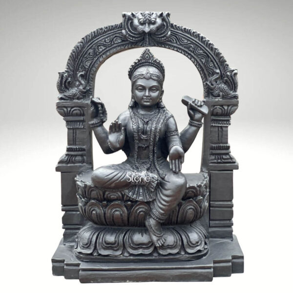 A full-frontal eye-level photograph of a meticulously hand-carved 2-foot Bala Tripura Sundari Black Stone Statue presented against a plain light gray background. The statue depicts the youthful Goddess Bala Tripura Sundari seated in a graceful lalitasana pose with her right leg folded and her left leg pendant resting upon a highly detailed multi-tiered lotus pedestal. She is carved from a single piece of dark polished black stone showcasing the natural texture and depth of the material. The goddess is adorned with intricate traditional jewelry including multiple necklaces armlets, bracelets and anklets as well as a finely carved crown and elaborate ear ornaments. Bala Tripura Sundari is depicted with four arms holding symbolic attributes. Her upper right hand holds an ankusha and her upper left hand holds a pasha. Her lower right hand is raised in the abhaya mudra conveying fearlessness and blessing while her lower left hand rests upon her knee holding a pustaka. Her face is serene and youthful with a gentle compassionate expression downcast eyes and a subtle smile reflecting deep spiritual calm and divine grace. The statue is framed by a beautifully carved prabhamandala which arches over the goddess's head and incorporates traditional South Indian temple architectural elements including stylized mythical creatures and floral patterns. This ornate arch is supported by two pillars that flank the seated figure enhancing the sense of a sacred shrine. The entire composition rests on a robust rectangular black stone base. The exquisite craftsmanship and fine detail of the carving highlight the spiritual significance and artistic beauty of this black stone statue of Bala Tripura Sundari.