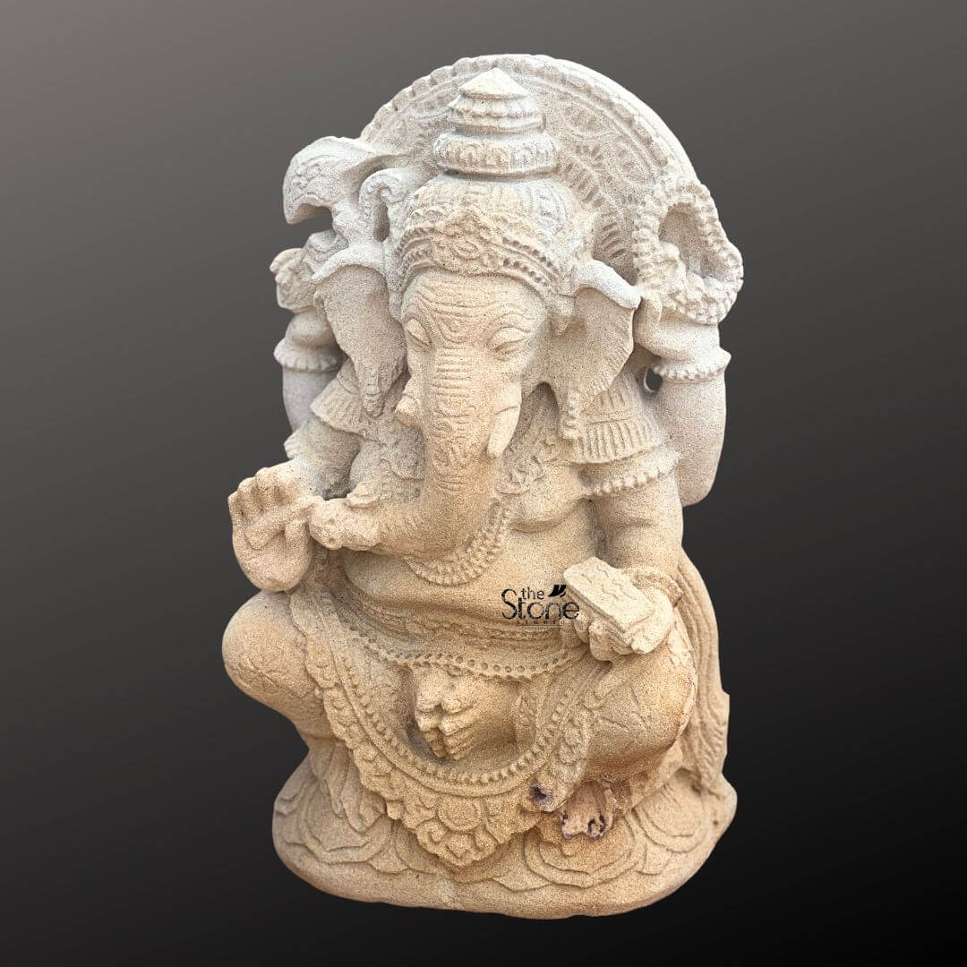 Indonesian Ganesha Idol for Home 2ft 2 A three-quarter view photograph of a magnificent 2-foot tall Bali Ganesha Statue meticulously hand-carved from warm beige sandstone presented against a dark neutral gray gradient background that accentuates its rich texture. The statue depicts the elephant-headed deity Lord Ganesha in a traditional seated posture upon a detailed lotus pedestal embodying profound tranquility and wisdom. The craftsmanship is highly detailed showcasing classic Balinese artistic influence with ornate carvings on his headdress jewelry and flowing garments. Ganesha is depicted with four arms holding symbolic attributes that convey his divine powers. His upper right hand holds an axe and his upper left hand holds a broken tusk while his lower right hand is raised in a gesture of blessing and his lower left hand holds a sweet that his trunk is tasting. The natural porosity and tonal variations of the sandstone are visible adding an earthy elegance and a sense of age to the piece. The statue's serene expression and detailed ornamentation make it a striking example of Balinese religious art. This 2-foot sandstone Ganesha statue is an ideal centerpiece for a home shrine meditation room or as a decorative element in a living space seeking a touch of Eastern spirituality and cultural heritage. Its compact size allows for prominent placement on an altar a console table or within a garden niche bringing a sense of auspiciousness and divine grace to its surroundings. The image clearly conveys the scale texture and intricate design of the idol making it a perfect visual reference for an article discussing Bali Ganesha statues.