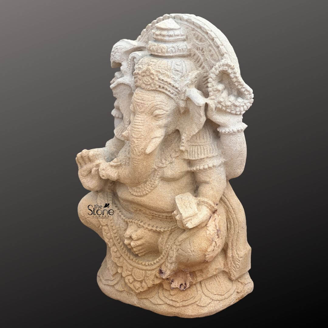 Indonesian Ganesha Idol for Home 2ft 6 A detailed view of a seated, antique-look Balinese Ganesha statue hand-carved from light beige sandstone. The statue depicts the elephant-headed Hindu deity Ganesha in a traditional Lalitasana pose with one leg folded and the other pendent resting on a detailed lotus-carved pedestal. Ganesha is adorned with intricate stylized Balinese-style jewelry including elaborate necklaces armbands and a richly carved crown that reflects the region's distinctive artistry. The sandstone surface shows a texture weathered patina, emphasizing the rustic and aged appearance requested. His main right hand is raised in the Abhaya mudra a gesture of fearlessness and blessing while his upper left hand holds an axe a symbol used in many Ganesha representations. His trunk gracefully curves towards a small bowl held in his lower left hand suggesting the offering of modak sweets. The sculptural details of the Balinese Ganesha statue are precise, showcasing the skill of the artisan in defining the folds of his garment the texture of his skin and the specific ornaments. The back of the statue features a decorative prabhavali framing his head and torso further enhancing its divine presence. The statue's posture and ornamentation are characteristic of Balinese Hindu iconography blending traditional elements with regional artistic expression. The sandstone has a natural color variation and porous texture giving it a authentic ancient feel suitable for the antique look described. This two-foot tall sandstone Ganesha statue is an excellent representation of Balinese stone carving art making it a relevant subject for the article on Balinese Ganesha statues. The seated posture the specific adornments and the overall antique finish capture the essence of traditional Balinese craftsmanship. Its size and detailed carvings make it suitable for both indoor and outdoor spiritual decor. The image clearly displays the unique style and cultural significance of this Balinese interpretation of Ganesha.