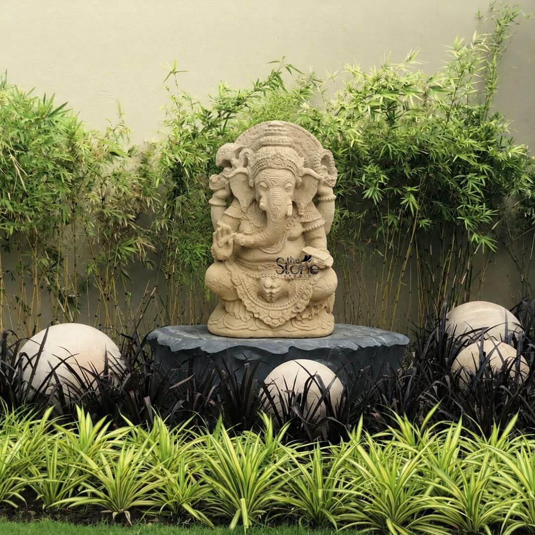 Indonesian Ganesha Idol for Home 2ft 3 A medium shot of a meticulously hand-carved antique-look Balinese Ganesha statue positioned centrally within a lush curated garden bed. The two-foot-tall statue hewn from light-beige sandstone depicts the elephant-headed Hindu deity Ganesha in a seated meditative pose known as Lalitasana with one leg folded and the other pendent. Ganesha is richly adorned with traditional Balinese jewelry including elaborate necklaces armbands and a textured crown all carved with deep relief that catches the natural light emphasizing the weathered aged patina of the stone. His trunk curves gently to his left, holding a sweet while his multiple hands bear symbolic attributes like an axe and a broken tusk rendered with fine detail. The entire form rests on a substantial dark polished stone pedestal which provides a strong contrast. The statue is set against a backdrop of vibrant, tall bamboo stalks that create a natural screen, their feathery green leaves adding depth and texture. In the foreground and surrounding the pedestal is a layered planting bed. The front edge is defined by low-growing variegated Chlorophytum comosum with green and white striped leaves. Behind these and flanking the statue's pedestal are dramatic dark-leaved plants possibly a variety of Ophiopogon planiscapus which further accentuates the light color of the Ganesha sculpture. Interspersed among the darker plants are several smooth light-grey river stones of varying sizes adding a natural sculptural element that balances the scene. This outdoor display beautifully exemplifies traditional Balinese stone sculpture as integrated into a modern landscape design. The antique finish of the sandstone gives the statue a sense of history and reverence characteristic of master craftwork from Bali. The composition highlights the statue's intricate details from the floral motifs on the crown to the pleats of the dhoti demonstrating the high level of skill in Balinese carving. The garden setting with its mix of tropical foliage and smooth stones provides a tranquil and appropriate context illustrating how such a sculpture serves as a focal point for meditation and aesthetic appreciation in a peaceful outdoor space. The image captures the spiritual and artistic essence of a Balinese Ganesha sculpture.