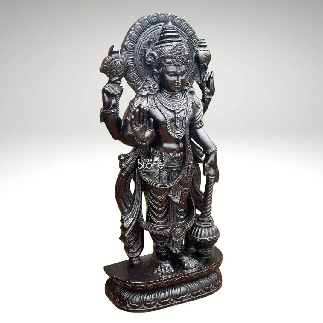 Black Stone Narayana Swamy Statue 3.5ft 2 A detailed full-body photograph captures a magnificent forty-one-inch-tall statue of Lord Narayana Swamy meticulously hand-carved from a single solid block of deep black granite. The deity is presented in a traditional standing posture upon an intricately carved circular lotus pedestal which features detailed petal layers and a decorative base. Narayana is depicted with four arms radiating divine grace and power making it an ideal sacred art piece for a home temple or place of worship. The statue exhibits a rich dark matte-to-satin finish characteristic of high-quality black granite which absorbs light and highlights the depth and complexity of the carvings. The four-armed form of Lord Narayana holds traditional symbolic attributes each intricately defined against the dark stone. His upper right hand holds the chakra while his upper left hand holds the shankha. His lower right hand is shown in the abhaya mudra a gesture of fearlessness and blessing and his lower left hand rests gracefully holding the gada. The deity is adorned with elaborate shallow-relief carvings of traditional South Indian temple jewelry including multiple necklaces armlets bracelets a sacred thread and a decorative mukuta all rendered with exceptional precision. The statue conveys a sense of profound spiritual calm and majesty with the intricate details of his flowing dhoti with elaborate folds the detailed garlands and the serene facial expression all contributing to its devotional appeal. The black granite material ensures durability and a timeless quality making this large statue a commanding presence. The statue is presented against a clean neutral graduated grey background ensuring the focus remains entirely on the detailed figure of Narayana Swamy and its artistic craftsmanship perfectly illustrating its suitability as a devotional focal point.