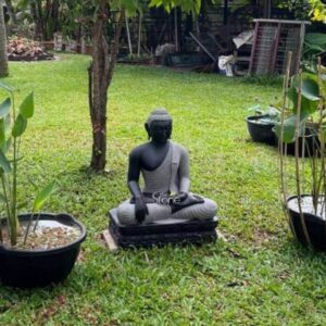 A striking 2-foot Black Stone Buddha statue ideal for a garden in a place like Kottayam Kerala is peacefully situated on a lush green lawn flanked by potted plants. The statue crafted with a distinctive two-tone finish – a dark black for the upper body and a contrasting lighter grey for the draped robes and lower body – depicts Buddha in a serene meditative posture seated in the full lotus position. This dual coloration emphasizes the contours and folds of the robes adding visual depth and artistry to the sculpture. His hands are in the Dhyana Mudra with both hands resting in his lap palms facing upwards and thumbs touching signifying meditation and inner peace. The Buddha's facial features are exquisitely carved radiating profound peace and wisdom with eyes gently closed or cast downwards in contemplation. His head is adorned with tightly coiled hair culminating in the ushnisha the cranial bump symbolizing his supreme spiritual knowledge. The unique coloration highlights the flowing drapery of his robes sculpted with soft elegant folds enhancing the statue's graceful and tranquil form. The statue rests on a beautifully sculpted lotus pedestal rendered in the dark black stone which in turn sits on a dark multi-tiered base providing a stable and dignified foundation. The Buddha is positioned centrally on a well-maintained green lawn with tall slender potted plants on either side adding to the natural serenity of the scene. The background reveals more greenery and trees suggesting a verdant possibly tropical garden environment typical of Kerala. This 2-foot Black Stone Buddha statue with its distinctive coloring and serene demeanor is an exquisite example of a garden deity designed to bring blessings peace and aesthetic beauty to any outdoor space perfectly aligning with an article focusing on such statues in Kottayam Kerala.