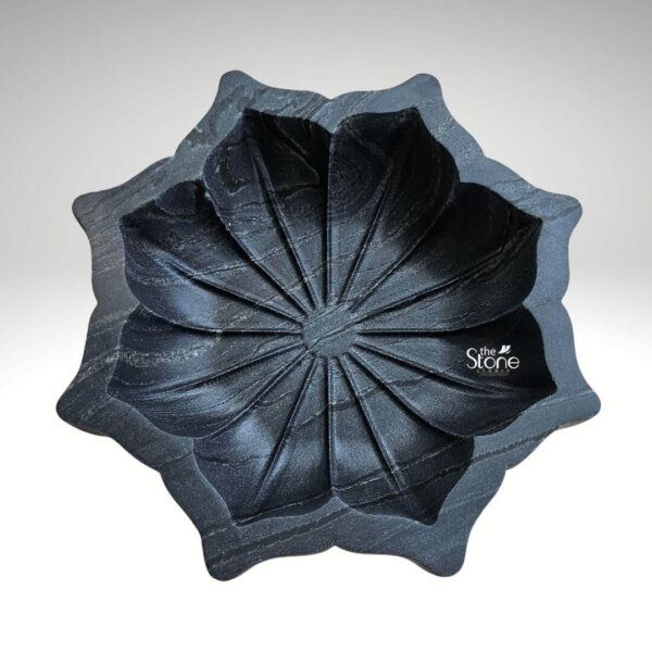 A high-angle overhead photograph displays a meticulously hand-carved black marble lotus-shaped Urli bowl measuring 12 inches in diameter. The bowl exhibits the deep rich black color and subtle greyish-white veining characteristic of premium black marble. It is perfectly centered against a clean neutral grey gradient background which isolates the object and emphasizes its form. The entire sculpture is presented as a single, cohesive piece of art showcasing the artisan's skill in transforming a solid block of stone into an intricate organic shape. The Urli bowl is sculpted to replicate a fully bloomed lotus flower with multiple layers of petals radiating outwards from a central core. The outer broader petals form the base and rim of the bowl while the inner more numerous petals define the interior basin. Each petal is carved with precision featuring soft rounded edges and gentle contours that create a sense of depth and natural realism. The surface of the marble is smooth and polished catching the light to highlight the detailed carving and the inherent beauty of the stone. This black marble lotus Urli statue is an exquisite example of traditional Indian craftsmanship and is a sought-after piece for home decor. The combination of the sacred lotus motif with the luxurious black marble makes it a striking focal point for any room whether placed on a console table used as a centerpiece with floating flowers and candles or integrated into a larger spiritual decor scheme. Its substantial 12-inch size ensures it makes a significant visual impact bringing a touch of elegance and tranquility to contemporary living spaces.
