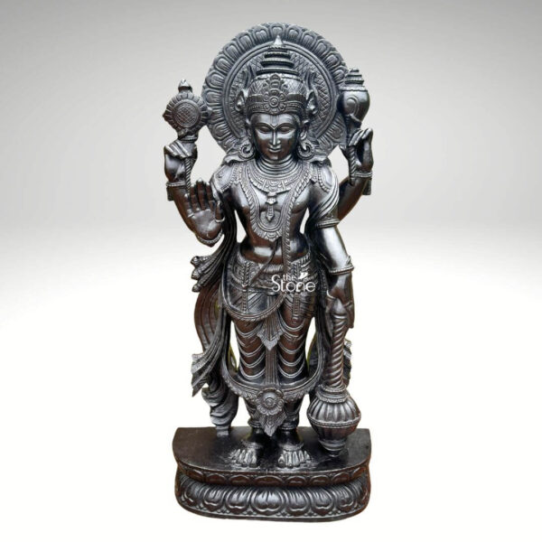A detailed full-body photograph captures a majestic forty-one-inch-tall statue of Lord Narayana Swamy meticulously hand-carved from a single solid block of deep black stone. The deity is presented in a traditional standing posture upon an intricately carved circular lotus pedestal which features detailed petal layers and a decorative base. Narayana is depicted with four arms radiating divine grace and power making it an ideal sacred art piece for a home temple or place of worship. The four-armed form of Lord Narayana holds traditional symbolic attributes each intricately defined against the dark polished stone. His upper right hand holds the chakra while his upper left hand holds the shankha. His lower right hand is shown in the abhaya mudra a gesture of fearlessness and blessing and his lower left hand rests gracefully holding the gada. The deity is adorned with elaborate shallow-relief carvings of traditional South Indian temple jewelry including multiple necklaces armlets bracelets a sacred thread and a decorative mukuta all rendered with exceptional precision. The statue exhibits a rich dark matte-to-satin finish characteristic of high-quality black stone which absorbs light and highlights the depth and complexity of the carvings. The intricate details of his flowing dhoti with elaborate folds the detailed garlands and the serene facial expression convey a sense of profound spiritual calm and majesty. The statue is presented against a clean neutral graduated grey background ensuring the focus remains entirely on the detailed figure of Narayana Swamy and its artistic craftsmanship perfectly illustrating its suitability as a devotional focal point. The base of the lotus pedestal is also carved with a textured pattern.