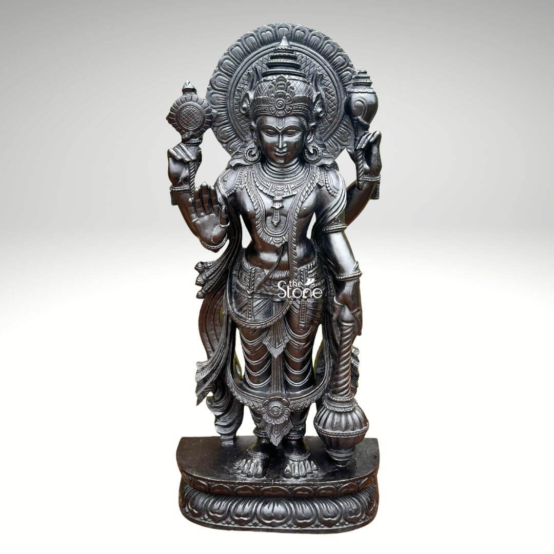 Black Stone Narayana Swamy Statue 3.5ft 1 A detailed full-body photograph captures a majestic forty-one-inch-tall statue of Lord Narayana Swamy meticulously hand-carved from a single solid block of deep black stone. The deity is presented in a traditional standing posture upon an intricately carved circular lotus pedestal which features detailed petal layers and a decorative base. Narayana is depicted with four arms radiating divine grace and power making it an ideal sacred art piece for a home temple or place of worship. The four-armed form of Lord Narayana holds traditional symbolic attributes each intricately defined against the dark polished stone. His upper right hand holds the chakra while his upper left hand holds the shankha. His lower right hand is shown in the abhaya mudra a gesture of fearlessness and blessing and his lower left hand rests gracefully holding the gada. The deity is adorned with elaborate shallow-relief carvings of traditional South Indian temple jewelry including multiple necklaces armlets bracelets a sacred thread and a decorative mukuta all rendered with exceptional precision. The statue exhibits a rich dark matte-to-satin finish characteristic of high-quality black stone which absorbs light and highlights the depth and complexity of the carvings. The intricate details of his flowing dhoti with elaborate folds the detailed garlands and the serene facial expression convey a sense of profound spiritual calm and majesty. The statue is presented against a clean neutral graduated grey background ensuring the focus remains entirely on the detailed figure of Narayana Swamy and its artistic craftsmanship perfectly illustrating its suitability as a devotional focal point. The base of the lotus pedestal is also carved with a textured pattern.
