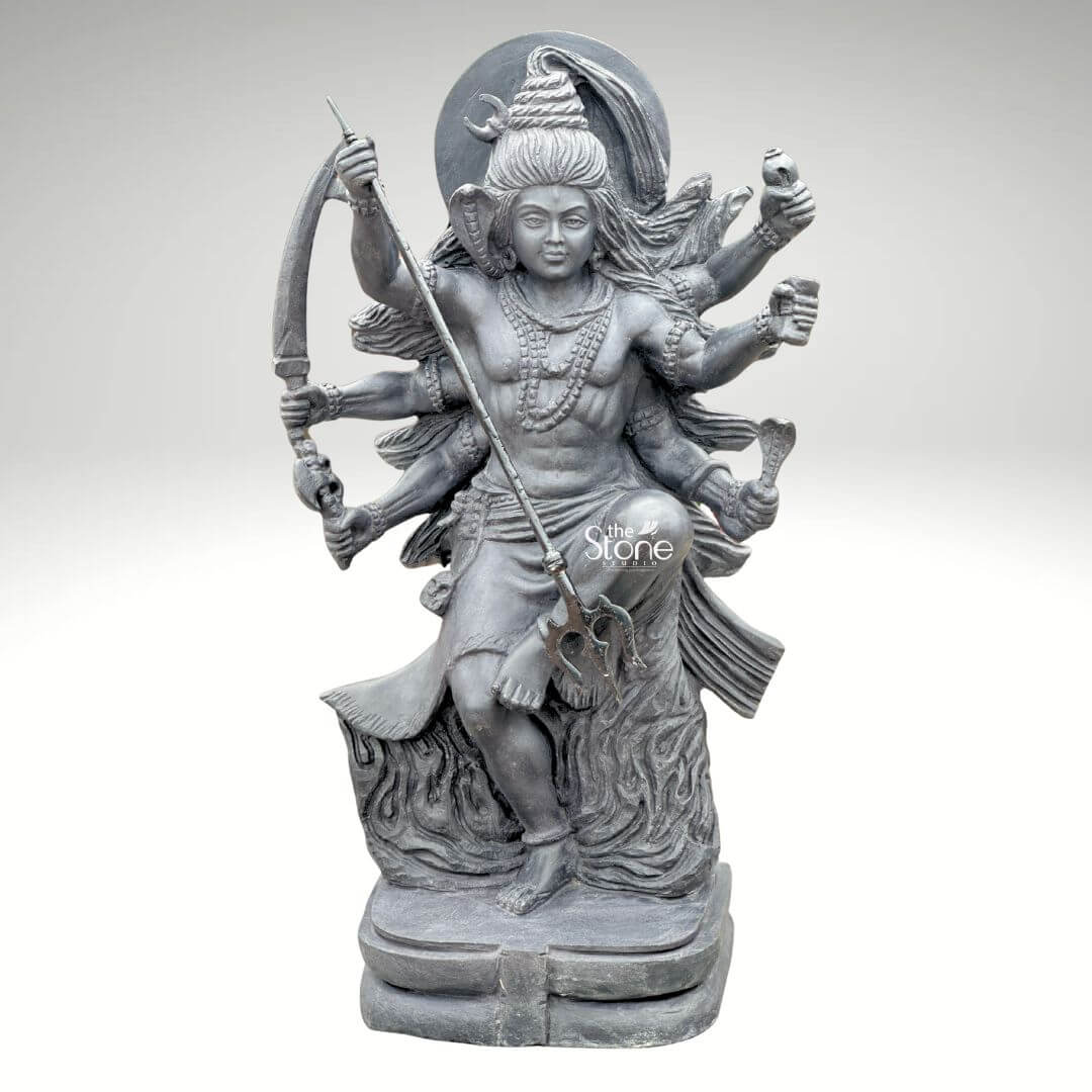 2 Feet Tandava Shiva Statue 5 A full-body studio photograph of a magnificent 2-foot tall Tandava Shiva idol masterfully hand-carved from a single block of dense polished black stone. The statue captures Lord Shiva in the dynamic and powerful Ananda Tandava pose the dance of bliss set upon a multi-tiered lotus pedestal. Shiva is depicted with ten arms fanned out to convey movement holding various divine attributes including a trident and damaru drum. His matted hair is piled high adorned with a crescent moon and a serpent and flares out dramatically to emphasize the energy of the dance. The intricate details of the carving from the folds of his garment to the expressions on his many faces are sharply defined showcasing the exquisite craftsmanship. The black stone used for the statue has a deep rich hue with a subtle waxy luster giving it a powerful and reverent presence. The ten arms are arranged with precision some raised in gestures of protection and blessing others holding attributes like a conch shell a fire pot and a scripture. Shiva's right leg is raised bent at the knee while his left leg is planted firmly on the demon Muyalakan who lies subdued beneath his foot. The dynamism of the dance is conveyed through the arching of his body and the intricate arrangement of his limbs creating a sense of controlled power and divine ecstasy. This 2-foot black stone Tandava Shiva idol stands as a testament to Indian temple art and a perfect centerpiece for any sacred space. Its compact size allows for placement on an altar or a display table where its intricate details can be admired. The dark monolithic stone gives it a timeless quality while the powerful iconography of the Tandava pose evokes a sense of cosmic energy and spiritual liberation. The photograph presents the idol against a clean neutral white background ensuring the focus remains entirely on the form texture and spiritual significance of the black stone sculpture.