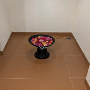 A centered eye-level photograph captures a traditional Indian Black Stone Urli placed as a decorative centerpiece in the corner of a room. The Urli a classic vessel used for floating flowers is hand-carved from a dark polished natural stone giving it a rich deep black color with subtle variations in texture. It features a wide shallow circular basin set atop a sturdy carved pedestal base elevating it off the brown tiled floor. The overall dimensions convey a substantial artisanal piece suitable for prominent placement. The basin of the Urli is filled with water creating a serene surface. Floating gracefully upon the water is a vibrant dense arrangement of fresh flowers. The predominant blooms are small multi-colored marigolds in shades of yellow and orange forming a lush bed of color. Nestled precisely in the center of this marigold arrangement is a single delicate open pink lotus flower serving as the focal point and providing a striking contrast to the dark stone and surrounding warm-toned petals. The flowers are fresh and appear carefully arranged. This elegant black stone Urli with its intricate floral display serves as a sophisticated and culturally rich element for home decor. The combination of the dark earthy stone and the bright natural colors of the flowers makes it a versatile piece that can enhance various interior styles from traditional Indian to contemporary minimalist. The presence of the lotus and marigolds adds a touch of auspiciousness and tranquility making it ideal for living rooms entryways or as part of a festive arrangement. The durable heavy stone construction ensures longevity establishing it as a permanent valuable addition to a home's decor.