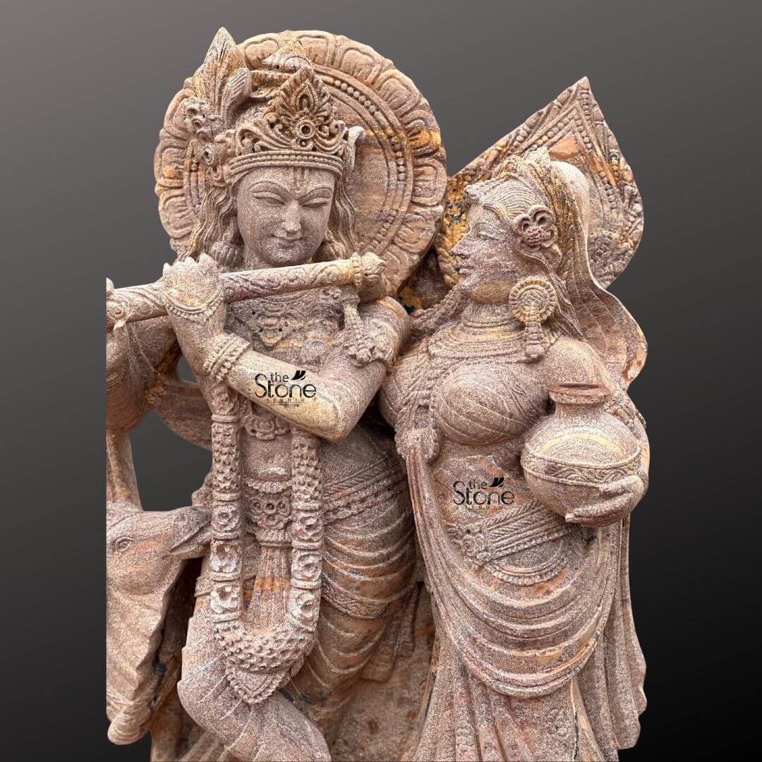 Radha Krishna Stone Statue with Cow 4ft 5 A close-up view of a magnificent 4-foot Brown Sandstone Radha Krishna Statue with Flute intricately carved from warm reddish-brown sandstone reveals the delicate artistry against a dark gradient background. The statue captures Lord Krishna and Radha in an intimate and loving standing pose. Krishna distinguished by his elaborate crown and serene expression holds a flute to his lips seemingly in the act of playing with his head gently tilted towards Radha. Radha stands gracefully beside him adorned with a similar crown and traditional attire her gaze fixed on Krishna while she carries a pot of water or butter in her hand. This detailed carving beautifully highlights their divine romance and interaction. The craftsmanship is exceptional with every intricate detail defining their elaborate crowns delicate jewelry and flowing traditional garments. The sandstone's natural texture and subtle variations in color enhance the antique feel of the sculpture bringing out the tender expressions on their faces and the intricate patterns of their attire. Krishna's halo-like circular aureole with ornate patterns is partially visible behind him emphasizing his divine presence. A small docile cow is subtly depicted near Krishna's feet symbolizing his role as a protector and caretaker of cattle adding to the pastoral charm. The figures stand on a multi-tiered intricately carved pedestal adorned with lotus petal designs and other traditional motifs providing a stable and dignified foundation. The depth and precision of the carving across the entire 4-foot statue are remarkable showcasing the artistry of the piece. This Brown Sandstone Radha Krishna statue with its prominent flute and tender depiction of the divine couple is an exquisite example of religious artistry designed to evoke feelings of love devotion and peace perfectly aligning with an article focusing on such charming and symbolically rich Radha Krishna statues.