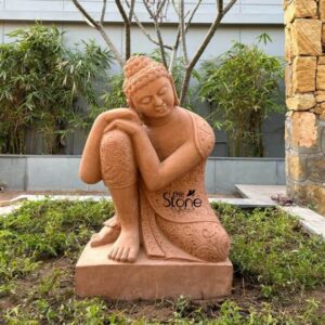 A full-shot outdoor photograph captures a beautifully sculpted three-feet tall sandstone Buddha garden statue resting upon a matching rectangular sandstone plinth. The statue is hand-carved from warm peach-colored sandstone exhibiting a natural slightly textured granular finish. It is positioned centrally within a modern garden landscape in Ahmedabad Gujarat nestled among low-lying green groundcover plants with a background of bamboo stalks and a contemporary building wall. The Buddha is depicted in a unique relaxed contemplative posture seated on the plinth with its left leg drawn up and right leg folded resting its head gently on its left knee with closed eyes conveying deep introspection. The statue displays intricate craftsmanship in its details. The Buddha's hair is carved in small tight traditional curls known as ushnisha typical of the style associated with stone carving in Rajasthan a common source for artisans in Gujarat. Its body is draped in a robe that features elaborate deep-relief carvings of floral and paisley motifs across the legs sleeves and hem showcasing the skill of the stonemason. The texture of the carved fabric contrasts with the smoother serene expression on the Buddha's face. This specific sandstone statue perfectly illustrates the fusion of traditional Indian artistry with contemporary garden design making it a sought-after element for serene outdoor spaces. The natural sunlight of Ahmedabad highlights the warm tones and the deep relief of the carvings enhancing its visual appeal. The statue is set against a backdrop of lush bamboo and a stone-clad pillar exemplifying how such a piece can serve as a focal point in a modern Indian garden.