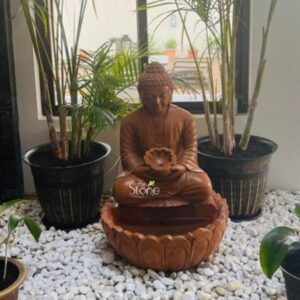 A medium shot captures a finely carved 3.5-foot-tall sandstone Buddha fountain statue positioned indoors against a large window overlooking an outdoor patio. The statue is hewn from warm reddish-brown sandstone displaying a natural slightly rough texture with visible grain and color variations giving it an authentic aged appearance. The Buddha is depicted in a seated meditative pose with legs crossed and hands resting in the lap in Dhyana mudra holding a small lotus-shaped basin from which water would gently flow. His face is serene with eyes half-closed in contemplation and a gentle smile characteristic of classical Buddhist art from the region. The traditional ushnisha and elongated earlobes are clearly defined and the folds of his monastic robe are rendered with soft flowing lines. The statue is elevated placed centrally within a circular lotus-shaped basin that forms the lower part of the fountain. This basin rests upon a bed of small smooth white river pebbles which covers the floor area and extends outwards to frame the base of the fountain creating a clean naturalistic foundation. Flanking the Buddha statue are two large dark grey fluted planters each holding a tall slender palm plant possibly a species of Dypsis lutescens which adds a touch of tropical greenery and verticality to the composition. The sunlight from the window illuminates the scene highlighting the warm tones and textures of the sandstone and the lush foliage casting gentle shadows that add depth. This 3.5-foot Sandstone Buddha Fountain statue serves as a perfect focal point for discussing stone decor elements suitable for tranquil spaces such as a home patio or interior courtyard in Botswana. The choice of durable sandstone reflects the region's appreciation for natural materials while the water feature adds a calming cooling element to the hot dry climate of Southern Africa. The statue's size is substantial enough to be a commanding presence yet intimate enough for a domestic setting. The image effectively showcases the harmonious blend of spiritual symbolism artistic craftsmanship and functional design making it an ideal visual reference for an article on incorporating such serene stone art into a home environment in Botswana.