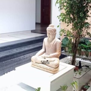 A serene two-foot-tall Buddha statue meticulously hand-carved from warm off-white sandstone sits in a traditional meditative pose on a raised white pedestal in the courtyard of a modern home in Kayamkulam Kerala. The statue depicts the Buddha with his legs crossed in a full lotus position hands resting gracefully in his lap in the dhyana mudra symbolizing deep contemplation and inner peace. His facial expression is calm and benevolent with downcast eyes a gentle smile and tightly curled hair leading to a pointed ushnisha all rendered with exquisite craftsmanship and refined flowing carved folds of his monastic robes. The setting is an artfully composed outdoor space that blends traditional sacred art with contemporary architecture. The Buddha statue is positioned on a clean white plinth making it a prominent focal point against the textured beige-to-grey wall behind it. To the right a lush tall tree and other smaller tropical plants add a vibrant touch of green anchoring the statue within the natural environment characteristic of Kerala. To the left a set of modern dark grey stone steps leads up to a brightly lit interior with white walls and a dark wooden door illustrating the integration of the statue into a residential living space. This photograph perfectly captures a beautiful installation featuring a 2 feet Buddha Off White Sandstone Statue making it an ideal illustration for an article discussing Buddha Stone Statue in Kayamkulam Kerala. The statue's natural stone texture harmonizes with the earthy tones of the surroundings while its calm presence brings a sense of tranquility to the modern home. The composition showcases the statue's role as a spiritual anchor and an aesthetic centerpiece demonstrating how such a piece can transform a simple patio or garden corner into a sacred and contemplative area. The detailed carving and thoughtful placement highlight its value as both a decorative and a meaningful addition to a Kayamkulam home.