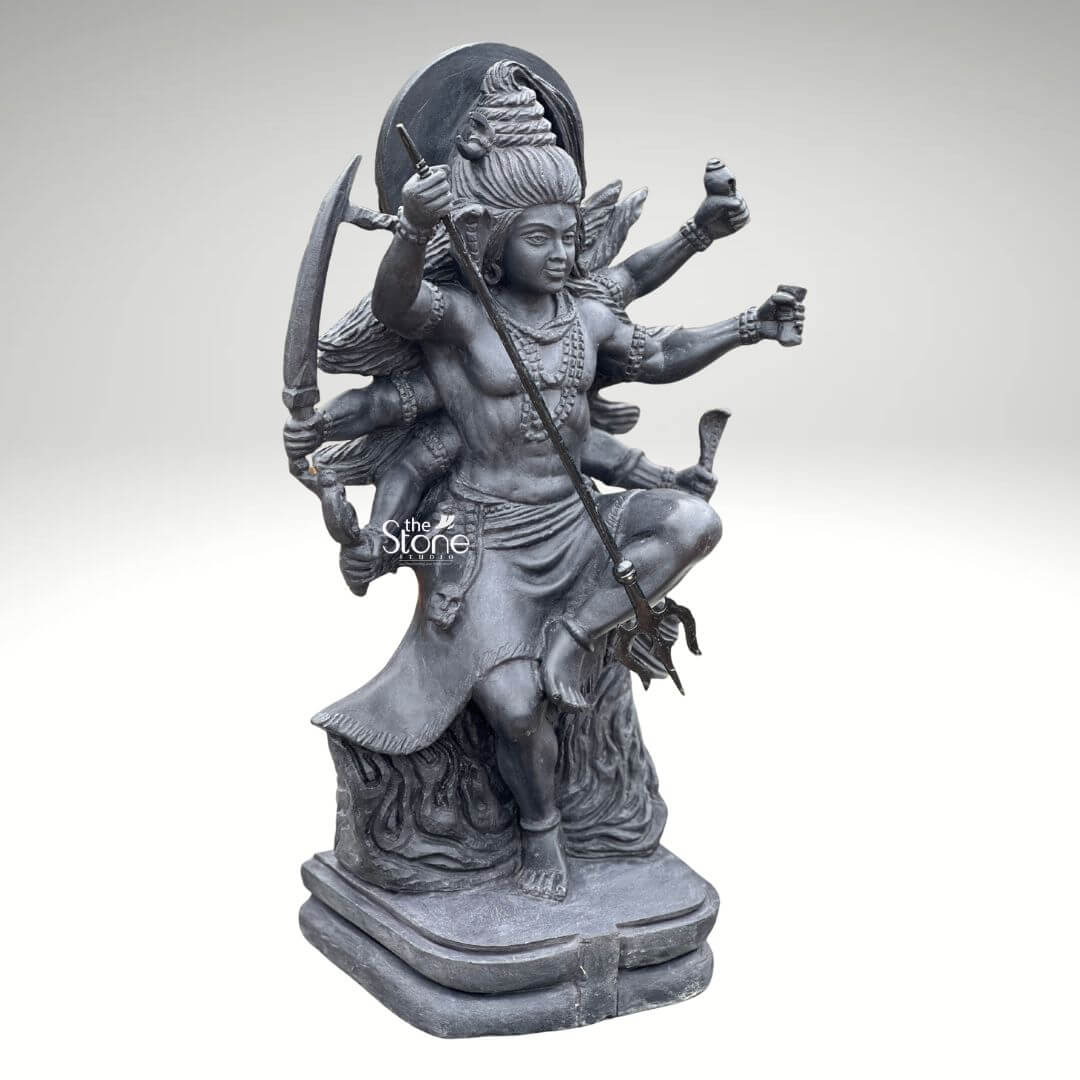 2 Feet Tandava Shiva Statue 6 A full-body studio photograph of a magnificent 2-foot tall Dancing Lord Shiva statue masterfully hand-carved from a single block of dense polished black stone. The statue captures Lord Shiva in the dynamic and powerful Ananda Tandava pose the dance of bliss set upon a multi-tiered lotus pedestal. Shiva is depicted with ten arms fanned out to convey movement holding various divine attributes including a trident and damaru drum. His matted hair is piled high adorned with a crescent moon and a serpent and flares out dramatically to emphasize the energy of the dance. The intricate details of the carving from the folds of his garment to the expressions on his many faces are sharply defined showcasing the exquisite craftsmanship. The black stone used for the statue has a deep rich hue with a subtle waxy luster giving it a powerful and reverent presence. The ten arms are arranged with precision some raised in gestures of protection and blessing others holding attributes like a conch shell a fire pot and a scripture. Shiva's right leg is raised bent at the knee while his left leg is planted firmly on the demon Muyalakan who lies subdued beneath his foot. The dynamism of the dance is conveyed through the arching of his body and the intricate arrangement of his limbs creating a sense of controlled power and divine ecstasy. This 2-foot black stone Dancing Lord Shiva statue stands as a testament to Indian temple art and a perfect centerpiece for any sacred space. Its compact size allows for placement on an altar or a display table where its intricate details can be admired. The dark monolithic stone gives it a timeless quality while the powerful iconography of the Tandava pose evokes a sense of cosmic energy and spiritual liberation. The photograph presents the statue against a clean neutral white background ensuring the focus remains entirely on the form texture and spiritual significance of the black stone sculpture.