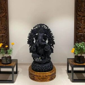 A 3.5 feet blackstone Ganesha statue is placed as the central focal point in a refined indoor setting creating a powerful and elegant visual presence. The statue depicts Lord Ganesha seated in a graceful posture on an ornately carved pedestal with the trunk gently curving downward and the body richly adorned with jewellery draped garments and intricate sculptural detailing. An elaborate decorative arch rises behind the figure framing the deity and adding depth grandeur and a temple-inspired character to the overall design. The deep black stone finish gives the Ganesha statue a bold timeless appearance while also highlighting the fine craftsmanship and detailed carving throughout the sculpture. The blackstone Ganesha statue is displayed on a circular carved wooden base that adds warmth and contrast to the dark stone. On both sides of the statue matching low wooden-and-metal side tables hold potted marigold plants, bringing a fresh touch of greenery and bright yellow-orange flowers to the composition. The arrangement is symmetrical and carefully balanced with tall carved wooden wall panels visible on the far left and right helping frame the central placement. The neutral cream wall and smooth flooring allow the decorative Ganesha statue to stand out clearly making the overall space look calm welcoming and thoughtfully styled. This image connects beautifully to an article about a Decorative Ganesha Statue in Mumbai by showing how a 3.5 feet blackstone Ganesha statue can transform a residential or interior décor setting into a space filled with spiritual charm and artistic sophistication. The placement demonstrates how such a statue can work well in an entrance foyer living area, lobby prayer corner or statement décor zone especially for homeowners looking for a premium devotional piece with strong visual impact. The combination of black stone detailed carving symmetrical styling floral accents and elegant surroundings makes this decorative Ganesha statue a striking example of luxurious home décor with cultural and spiritual significance.