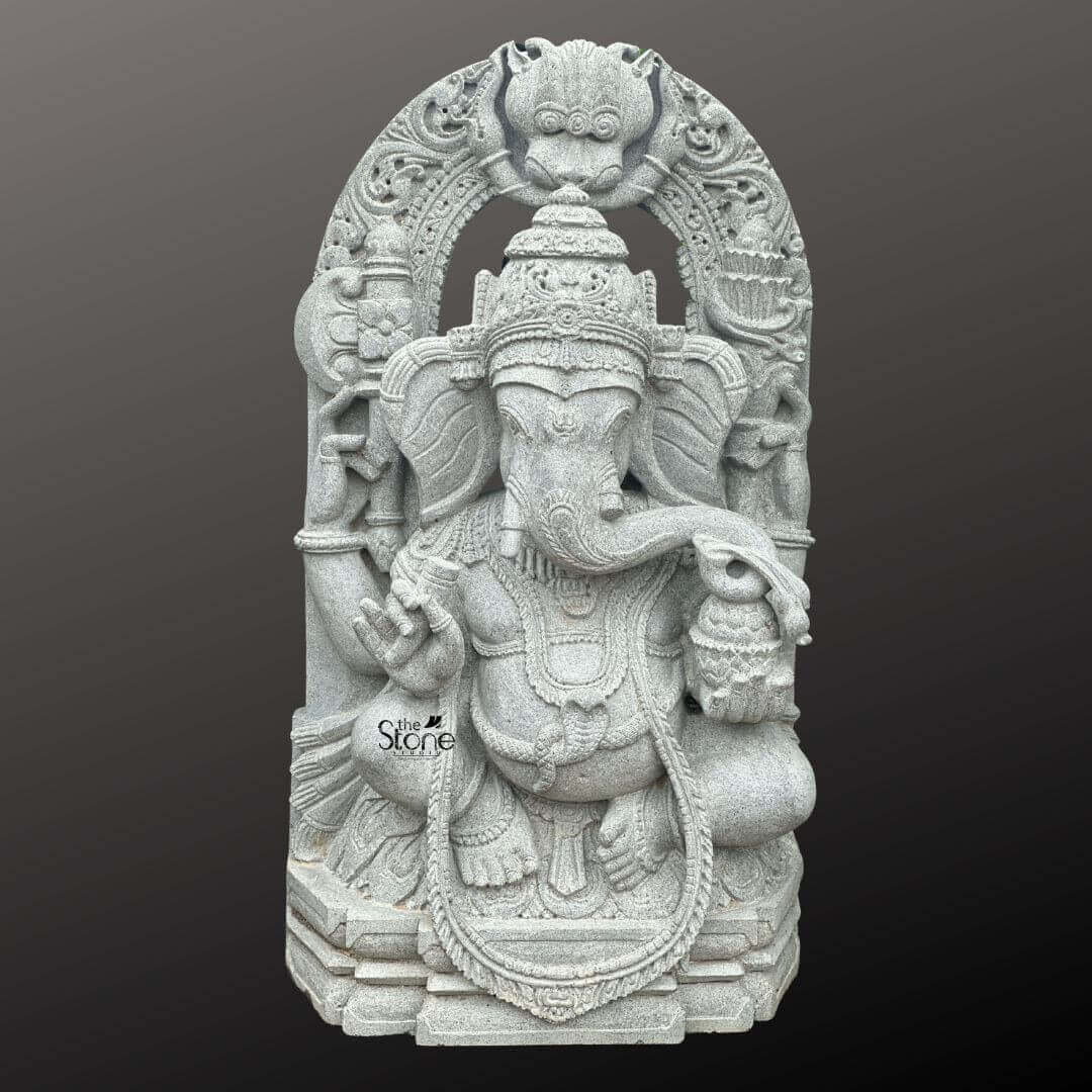 Ganesha Statue with Makara Torana 3ft 1 A magnificent 3-foot Ganesha statue with Makara Torana intricately carved from what appears to be a light grey stone stands powerfully against a dark gradient background. The statue depicts Lord Ganesha in a regal seated posture exuding benevolence and wisdom. His iconic elephant head is finely sculpted complete with large ears expressive eyes and his distinctive broken tusk. He is adorned with an elaborate crown multiple layers of traditional jewelry including necklaces armlets and a sacred threa draped across his chest all rendered with exceptional detail in the light grey stone. Ganesha is portrayed with four arms holding his customary attributes: a modak in one hand an axe or goad in another and his lower right hand extended in a gesture of blessing offering reassurance and protection. The intricate details on his garments the patterns on his halo and the careful sculpting of his pot belly are all clearly visible showcasing the artistry of the carving. The light grey stone emphasizes these textures and details lending a serene and timeless elegance to the deity. The statue is dramatically framed by a grand ornate archway clearly a Makara Torana which features exquisite mythical sea-creature motifs at its base and intricate traditional carvings along its entire span and supporting pillars. The top of the arch features a Kirtimukha a fierce lion-like face a common protective motif. This entire elaborate structure rests on a multi-tiered base also carved from the same light grey stone providing stability and enhancing its grand temple-like presence. This 3-foot Ganesha statue with its impressive Makara Torana is an exquisite example of religious artistry designed to bring blessings peace and aesthetic grandeur to any space perfectly aligning with an article focusing on such a revered Ganesha sculpture.